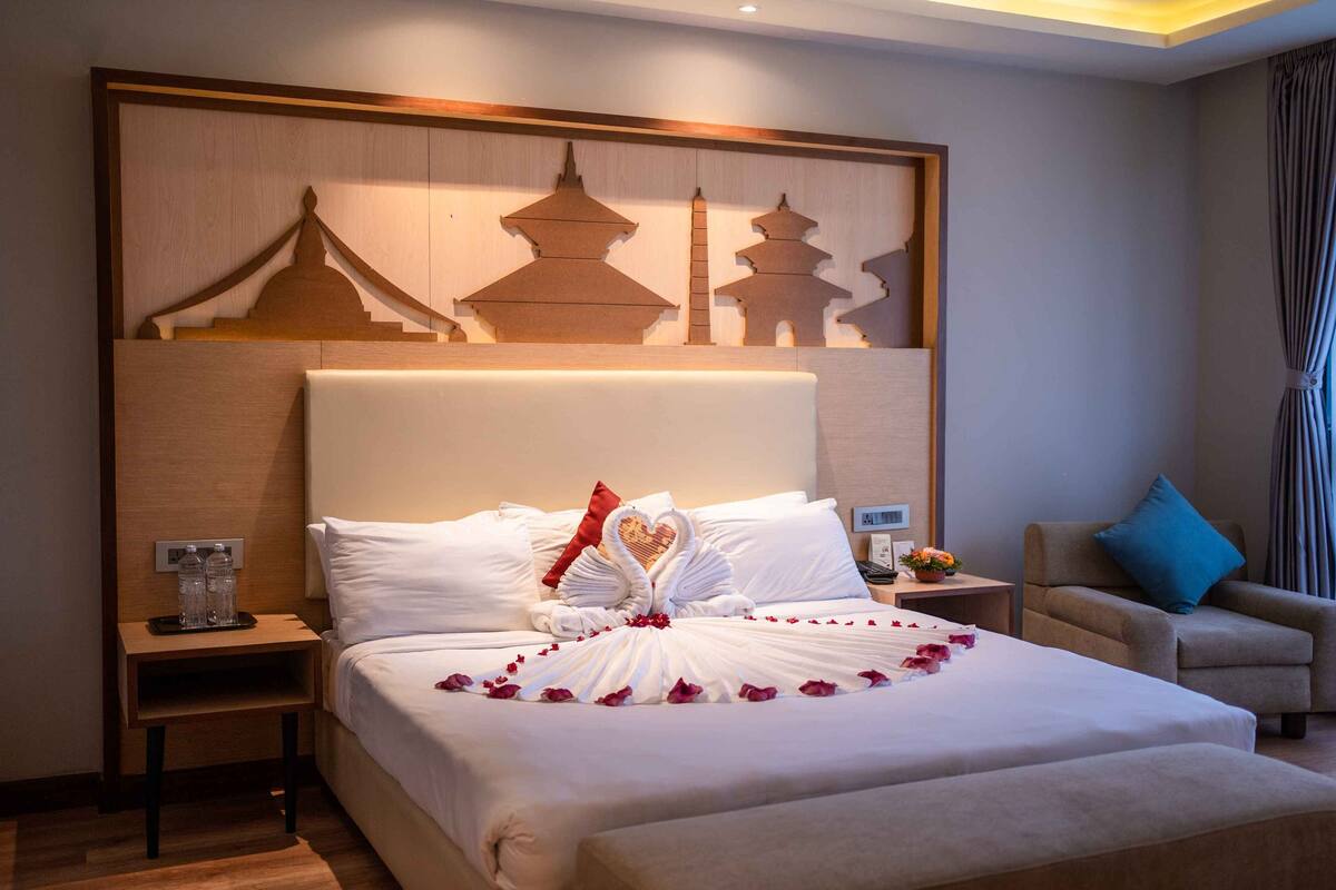 A hotel room with a large bed, a nightstand with a bottle and glass, a chair, and a wall with a decorative panel.