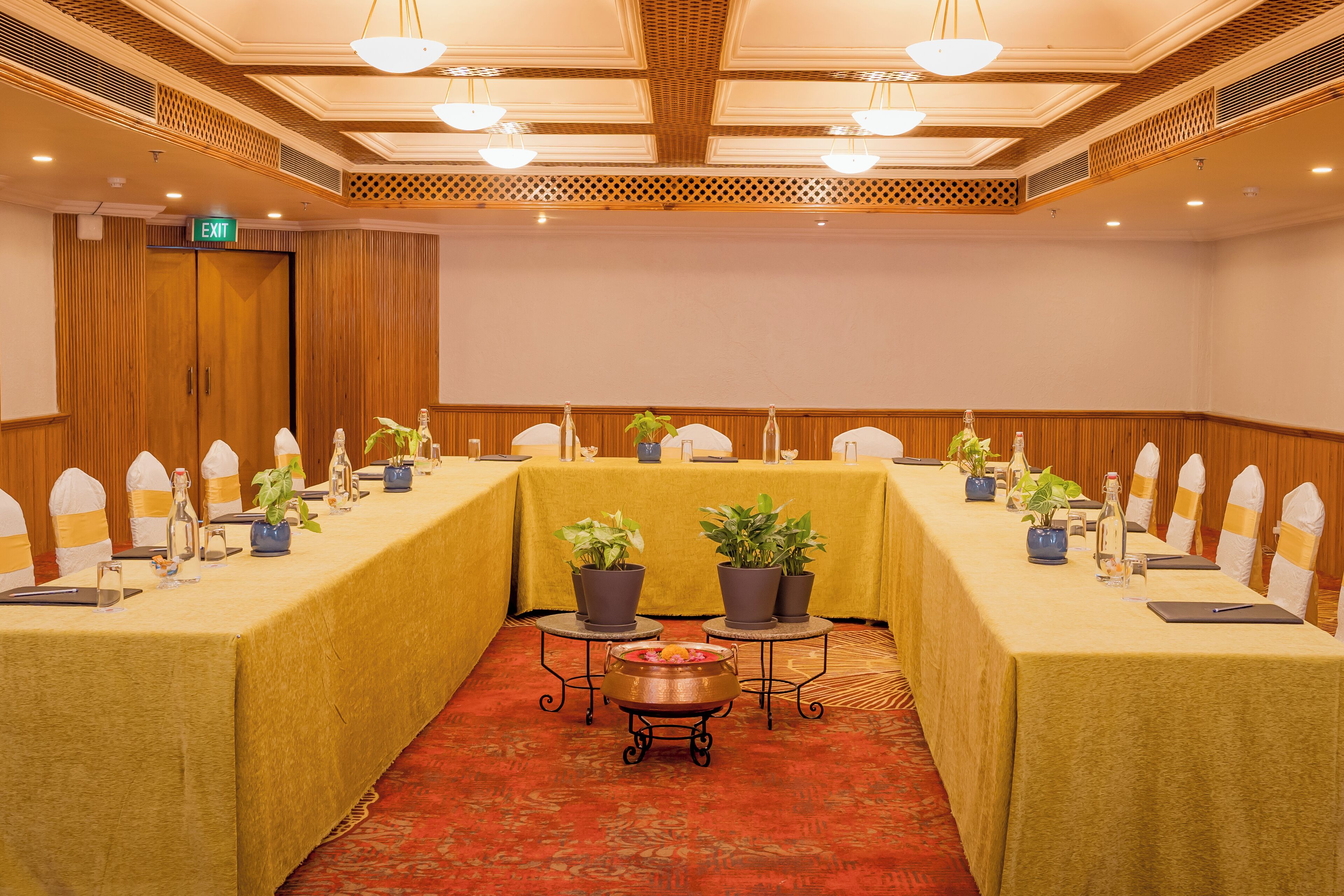 Una sala de conferencias con una mesa larga, sillas, plantas en macetas y una pequeña mesa con un centro de mesa decorativo.