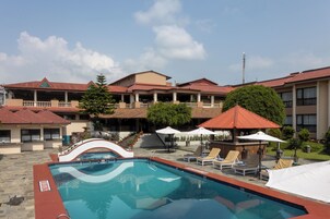 Una piscina con sillones de descanso y sombrillas, rodeada por un edificio con varios balcones y un techo de tejas rojas.
