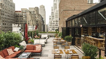 Um terraço no telhado com área de estar externa, bar e vista para os edifícios ao redor.