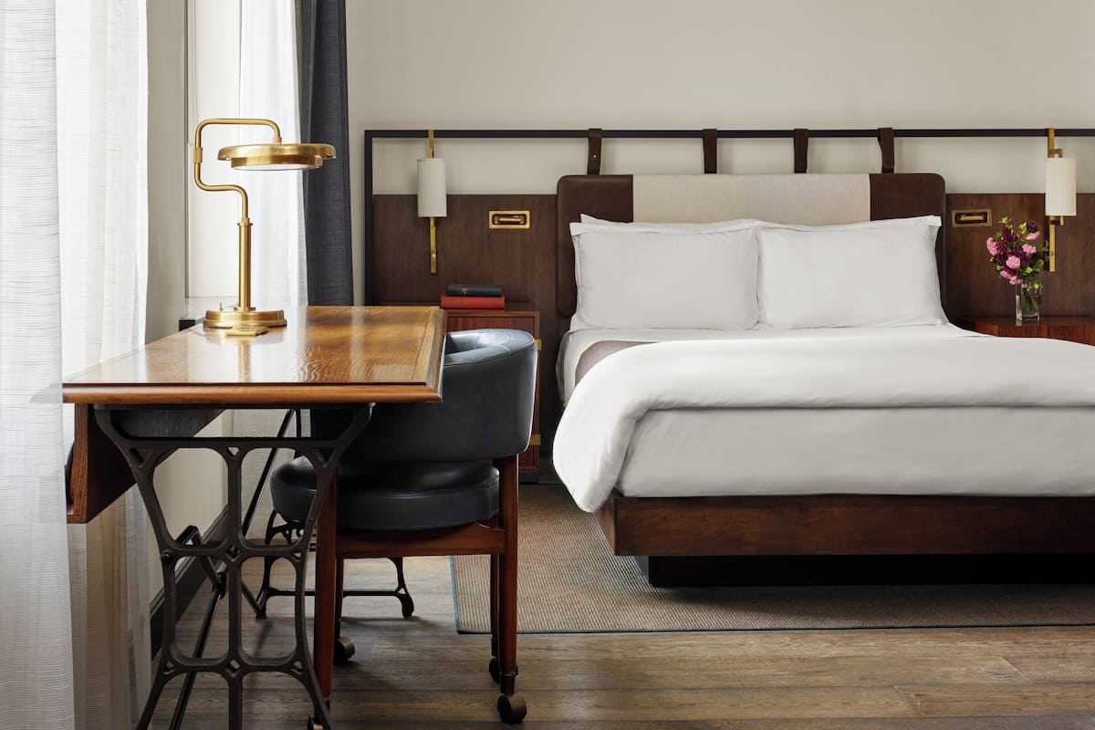 Ein Hotelzimmer mit Holztisch, Stuhl, Bett mit weißer Bettwäsche und Nachttisch mit Lampe.