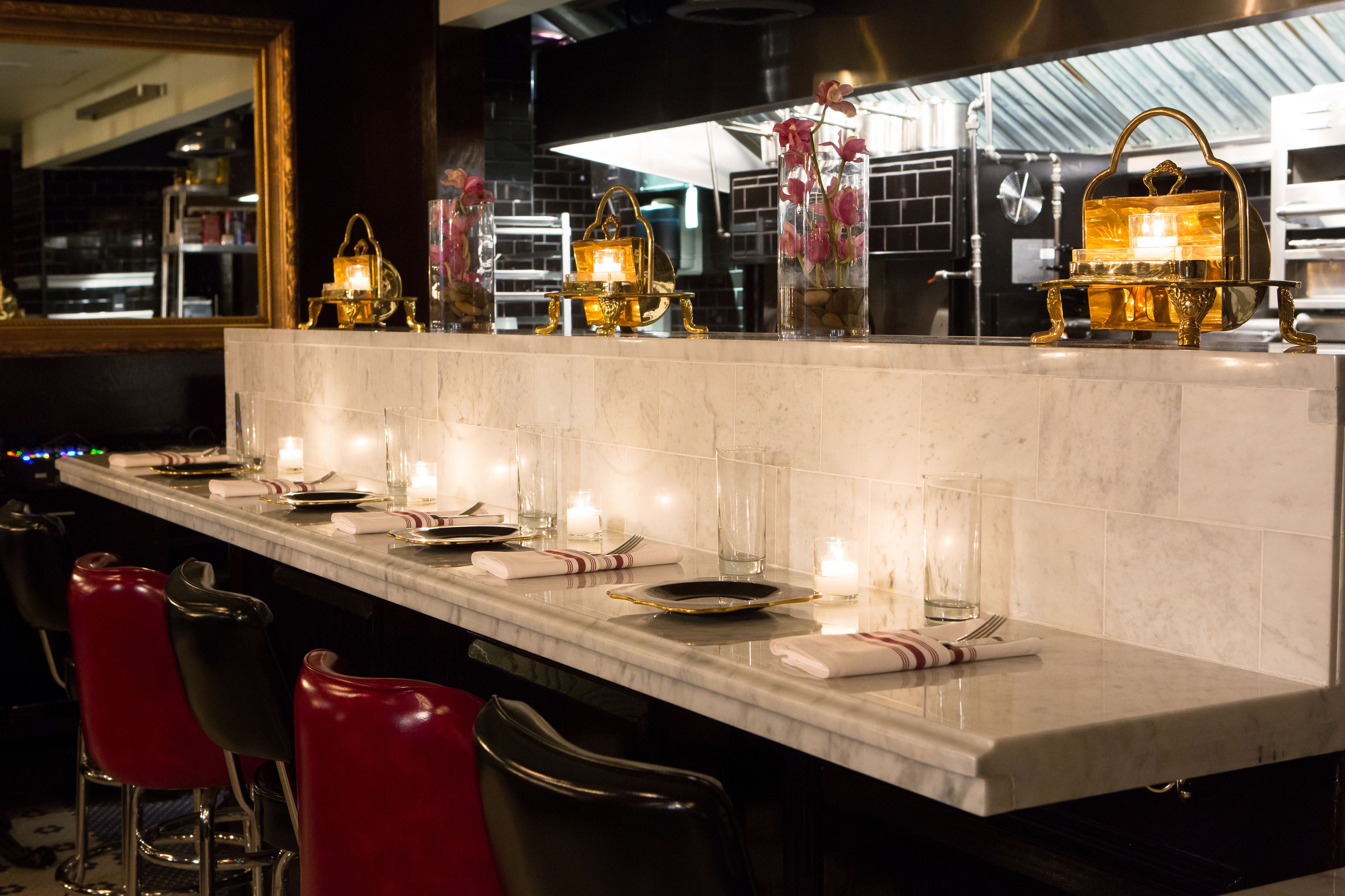 Una zona de bar con mostrador de mármol, taburetes y elementos decorativos como velas y recipientes de vidrio.