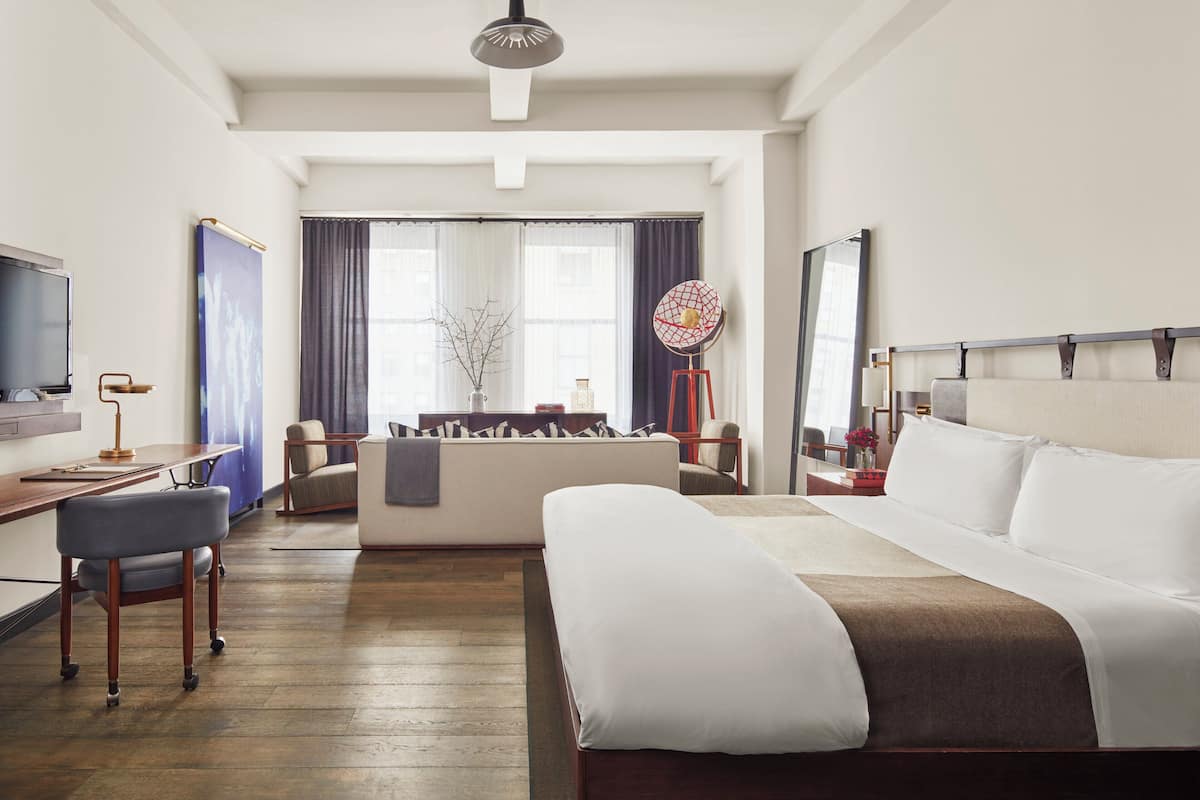 Ein Hotelzimmer mit einem großen Bett, einem Schreibtisch mit Computer, einem Sessel, einer Couch, einem Fernseher und einem Fenster mit Vorhängen.