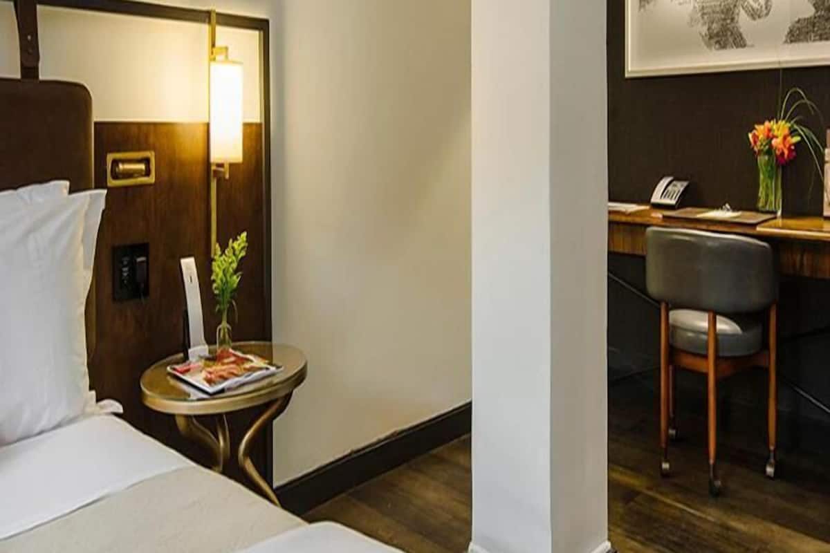 Ein Hotelzimmer mit Bett, Nachttisch mit Lampe, kleinem Schreibtisch mit Stuhl und Blumen in einer Vase.