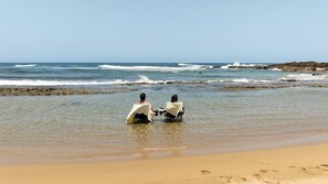 Two people sitting in chairs at the edge of a beach with waves in the background.