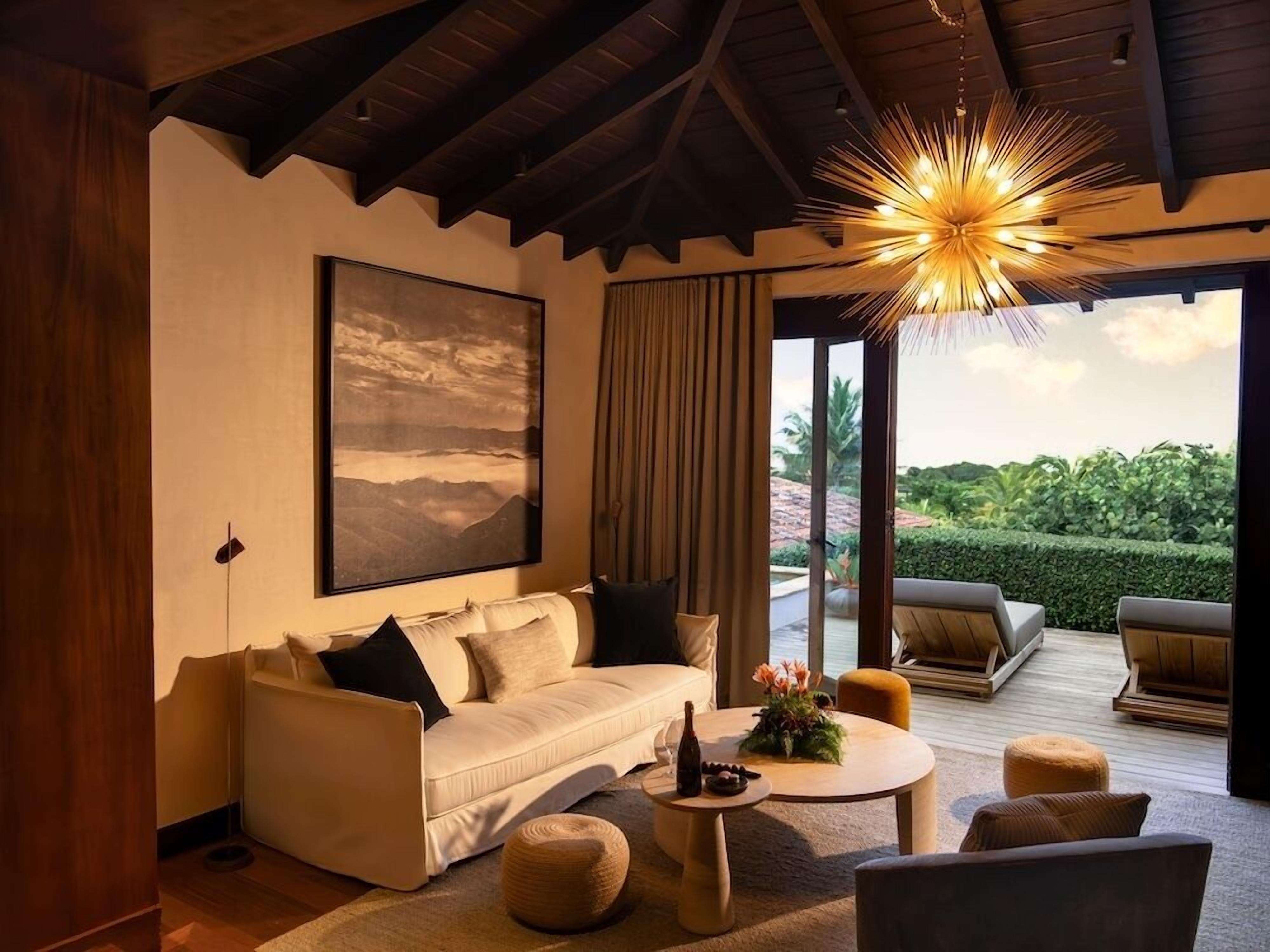A modern living room with a sofa, coffee table, and chairs, a large painting, and a view of greenery outside.