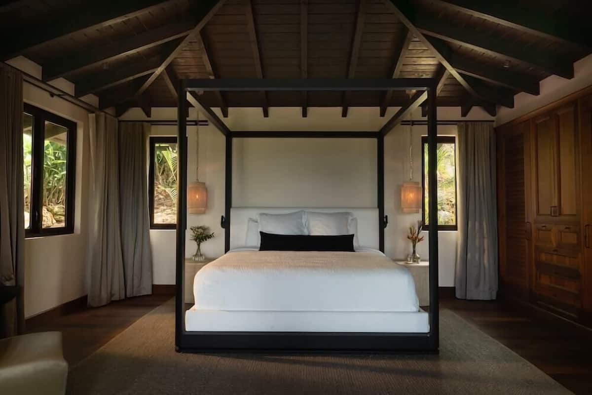 A four-poster bed in a room with wooden beams on the ceiling, large windows, and a nightstand with a lamp.