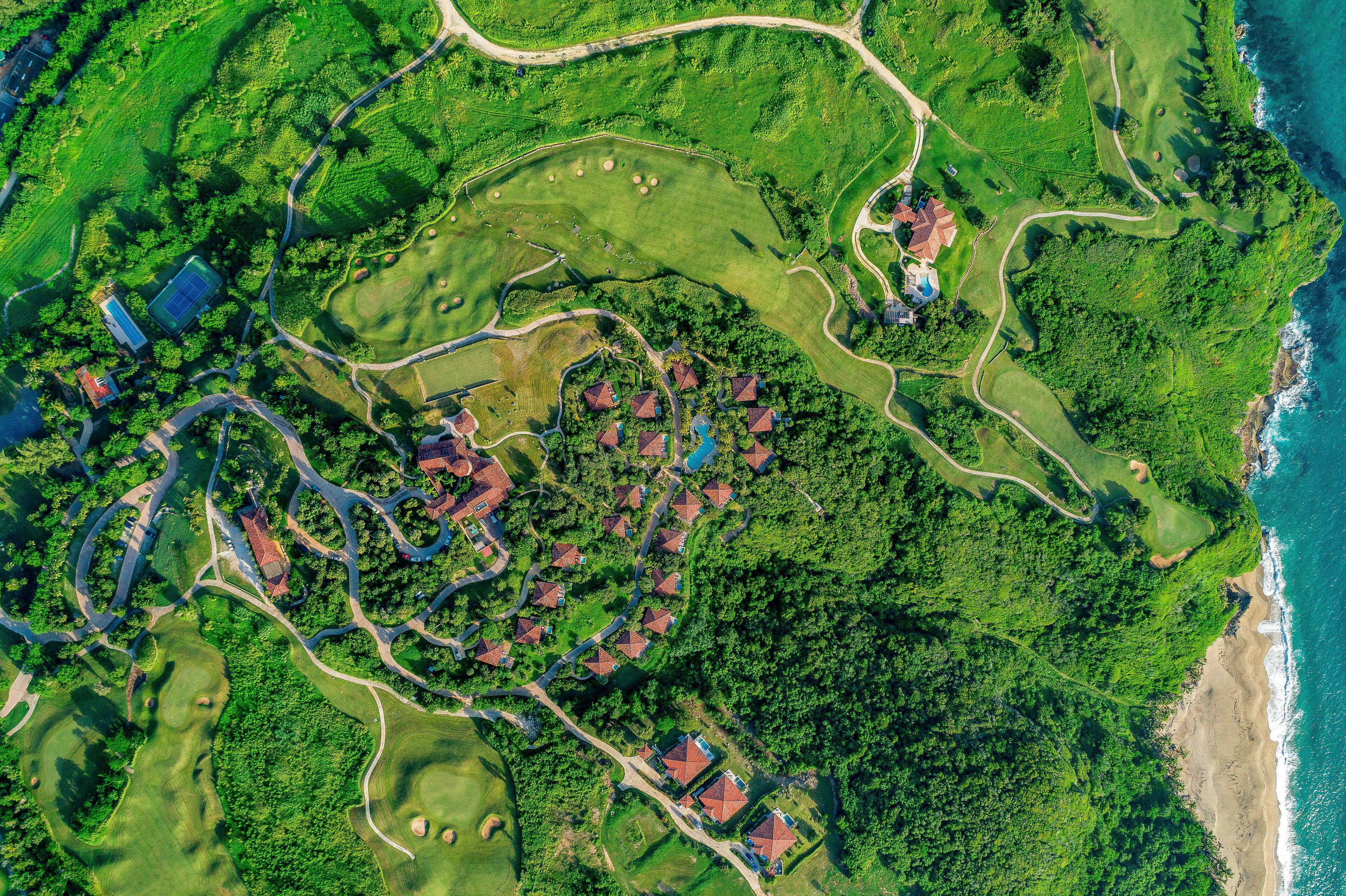 An aerial view of a golf course with multiple bunkers, a winding road, and several buildings with red roofs.
