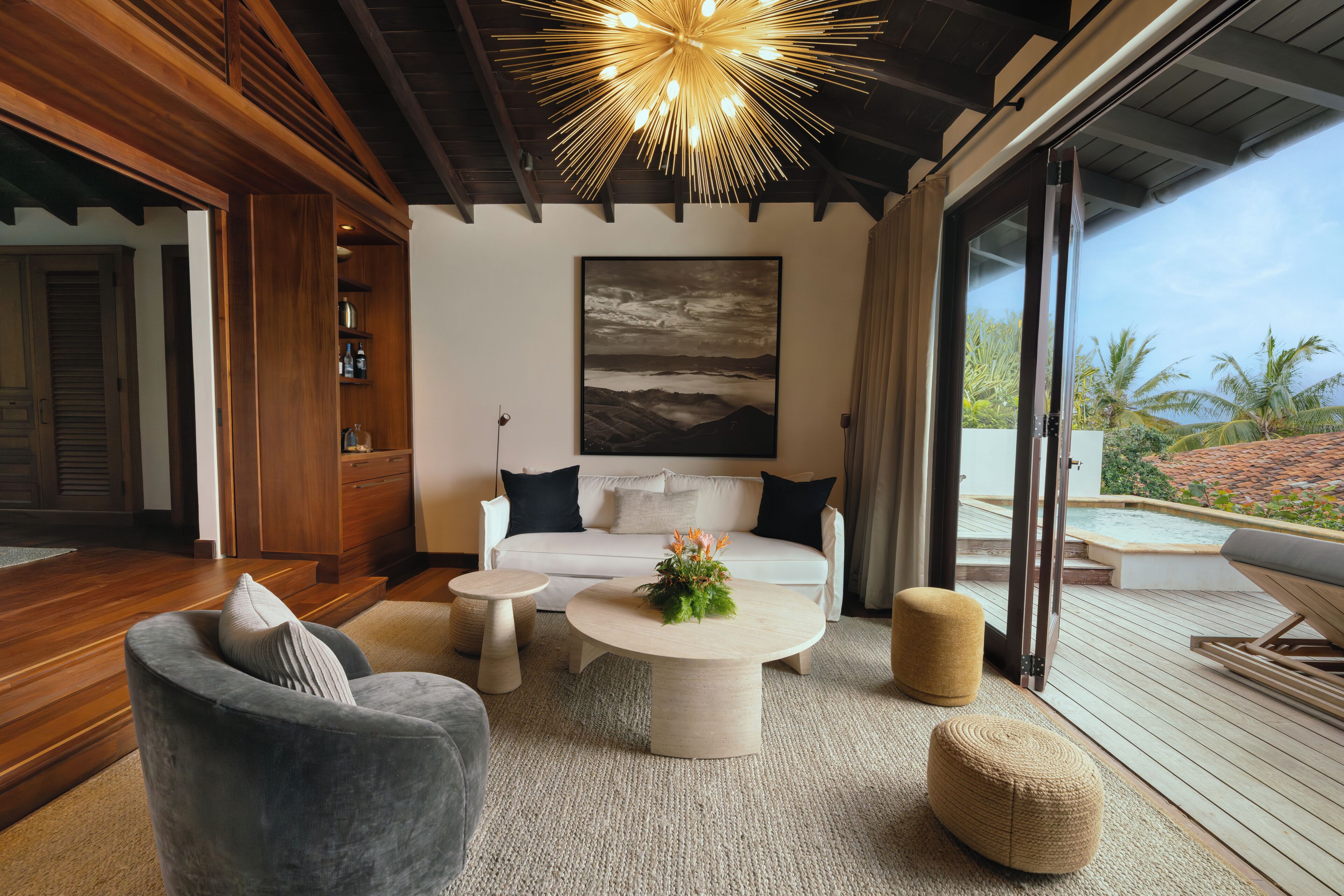 A modern living room with a white sofa, a round coffee table, and a grey armchair. There is a large painting on the wall and a view of the outdoors through a sliding glass door.