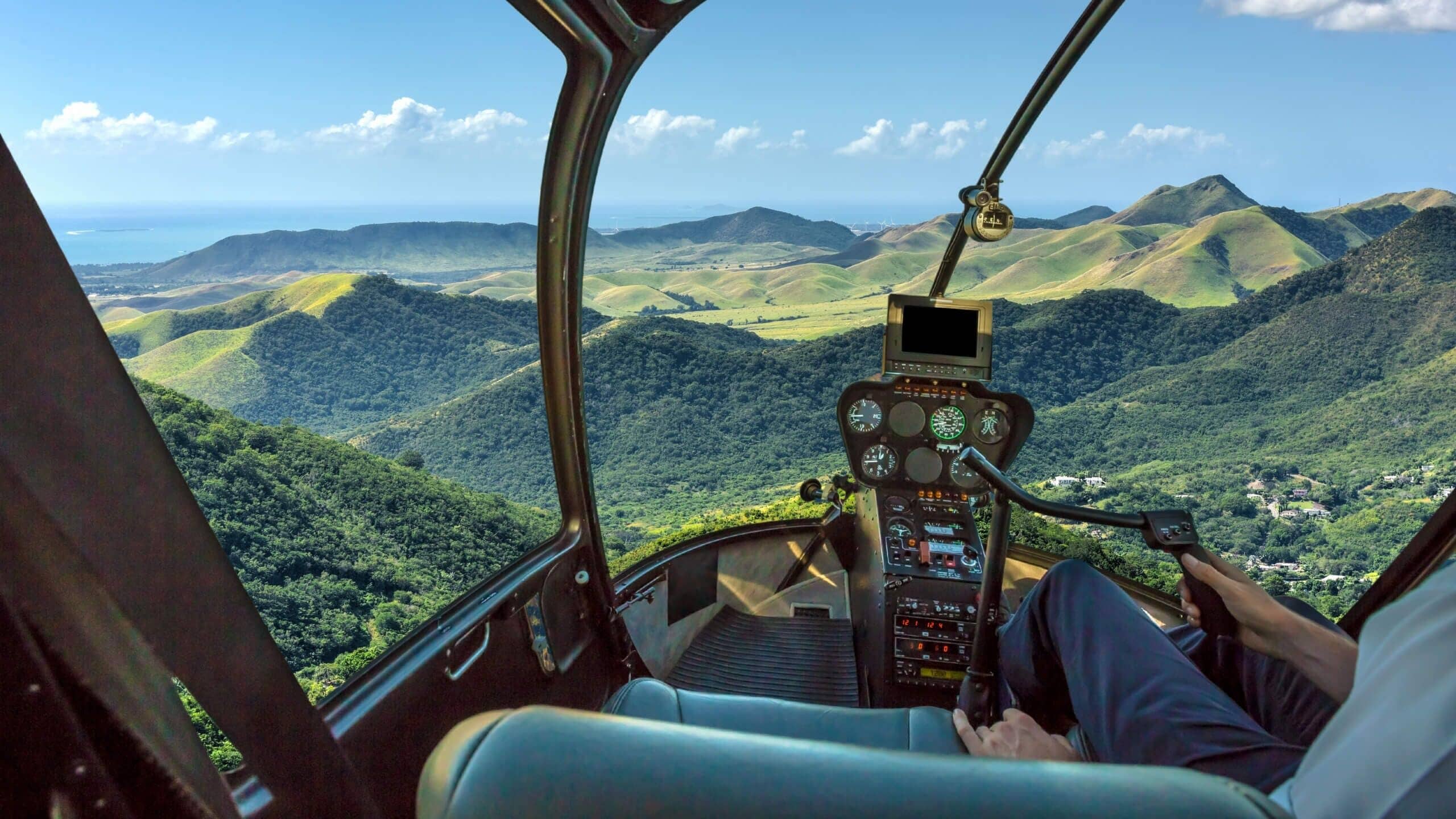 Helikoptercockpit met uitzicht op groene heuvels en een verre waterpartij.
