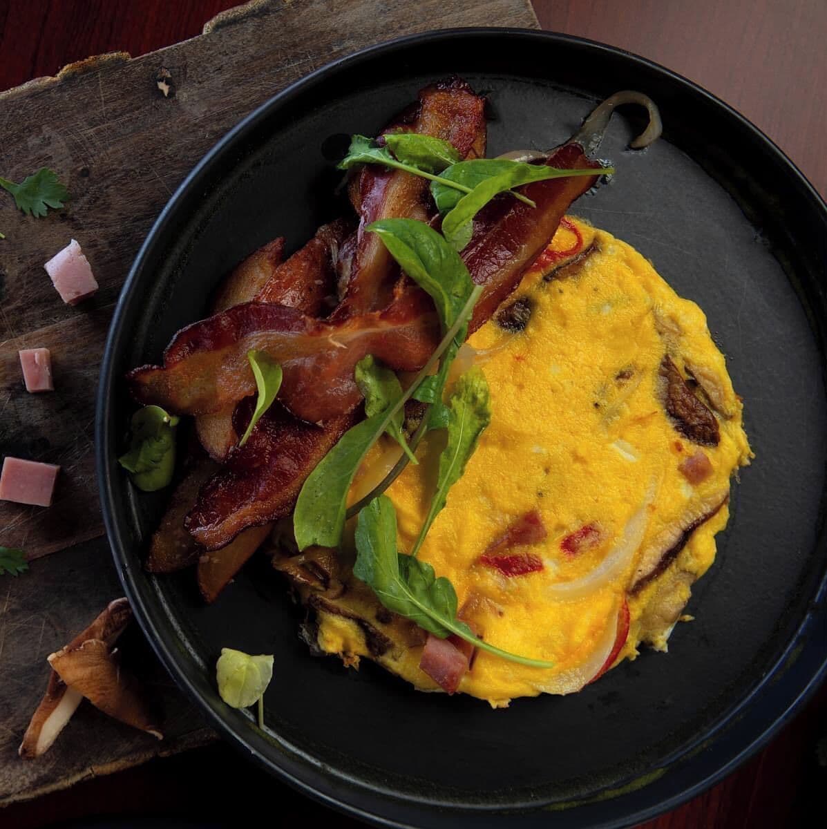 A plate with a fried egg, bacon, and greens on a dark plate.