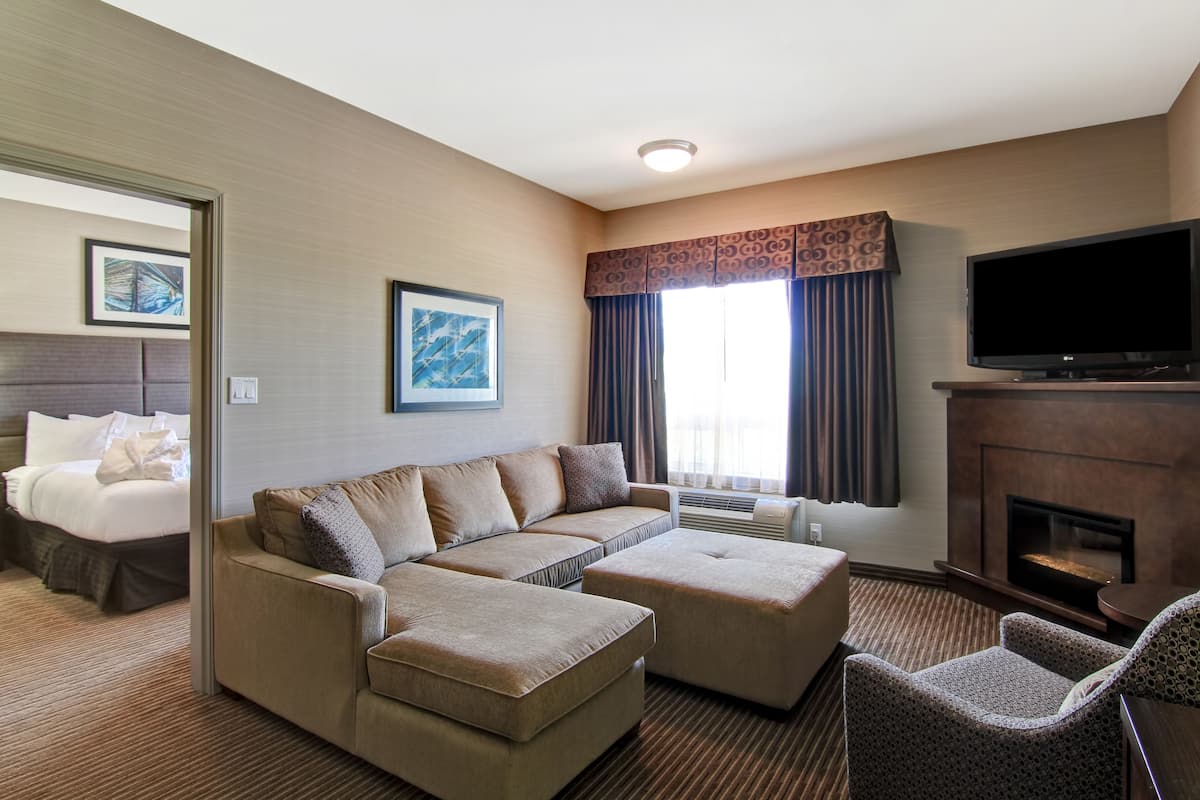 A hotel room with a sectional sofa, a flat-screen TV, a fireplace, and a bed with a headboard.