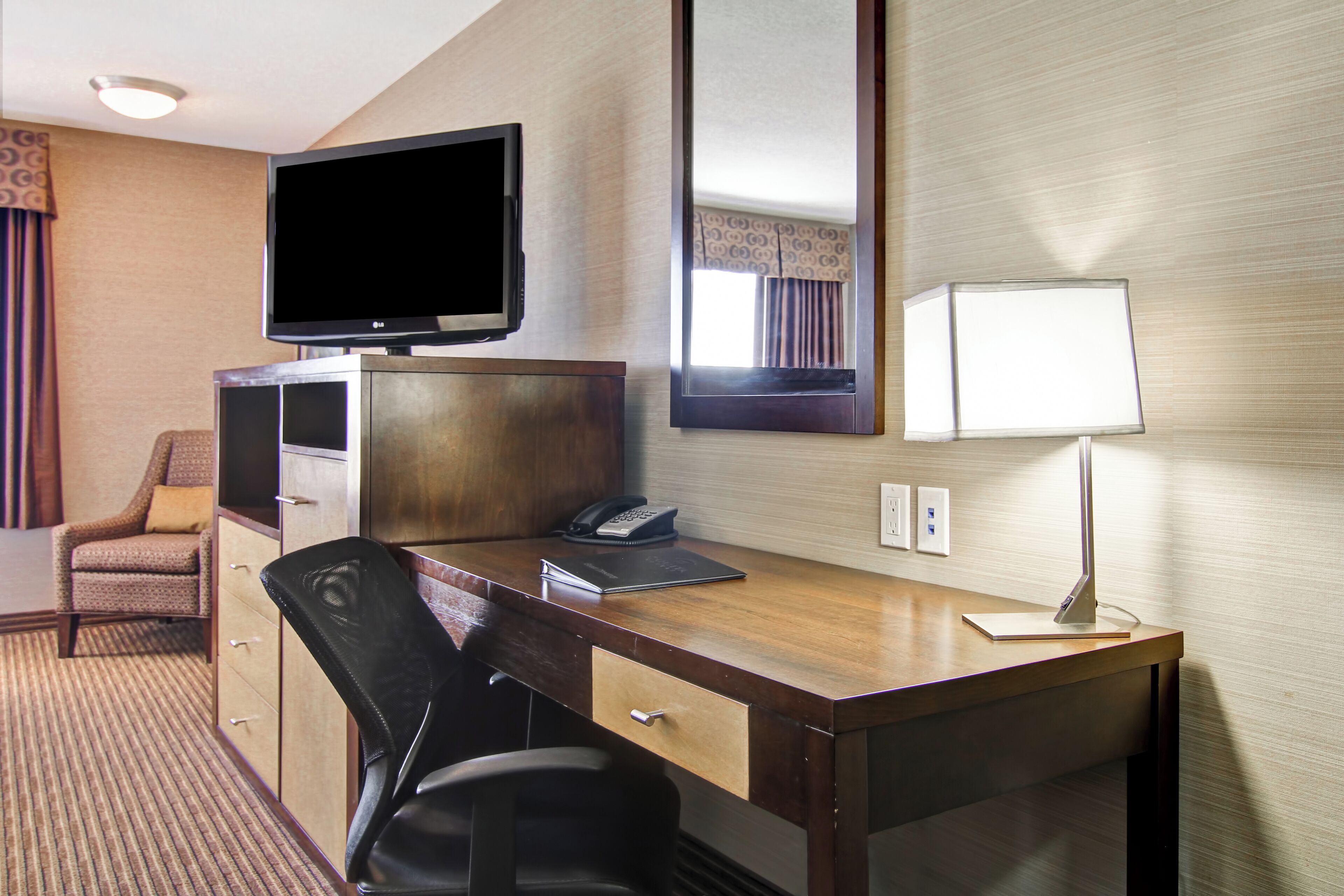 Habitación de hotel con un escritorio de madera, televisión, lámpara, silla, teléfono y espejo.