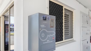 A KeySafe device mounted on a wall next to a window with a security grille. - Citywalk Motor Inn (Rockhampton)