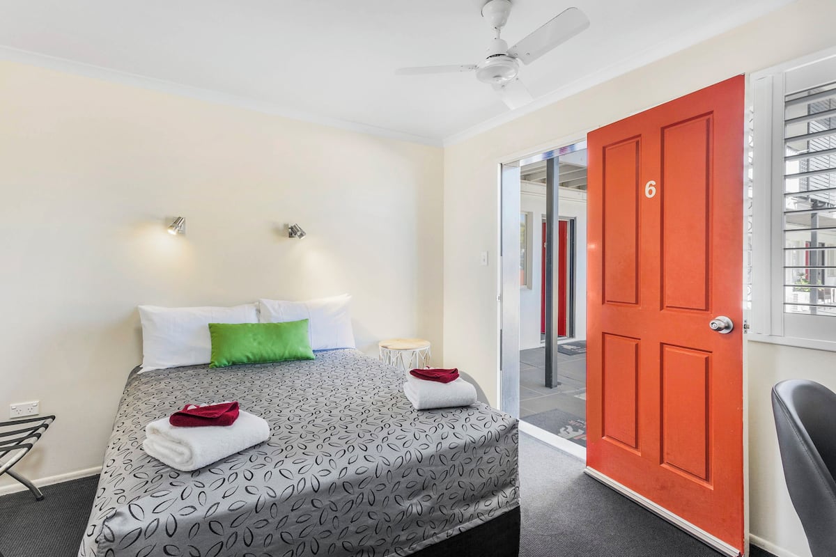 Habitación de hotel con cama, puerta roja, ventilador de techo y una pequeña mesa.