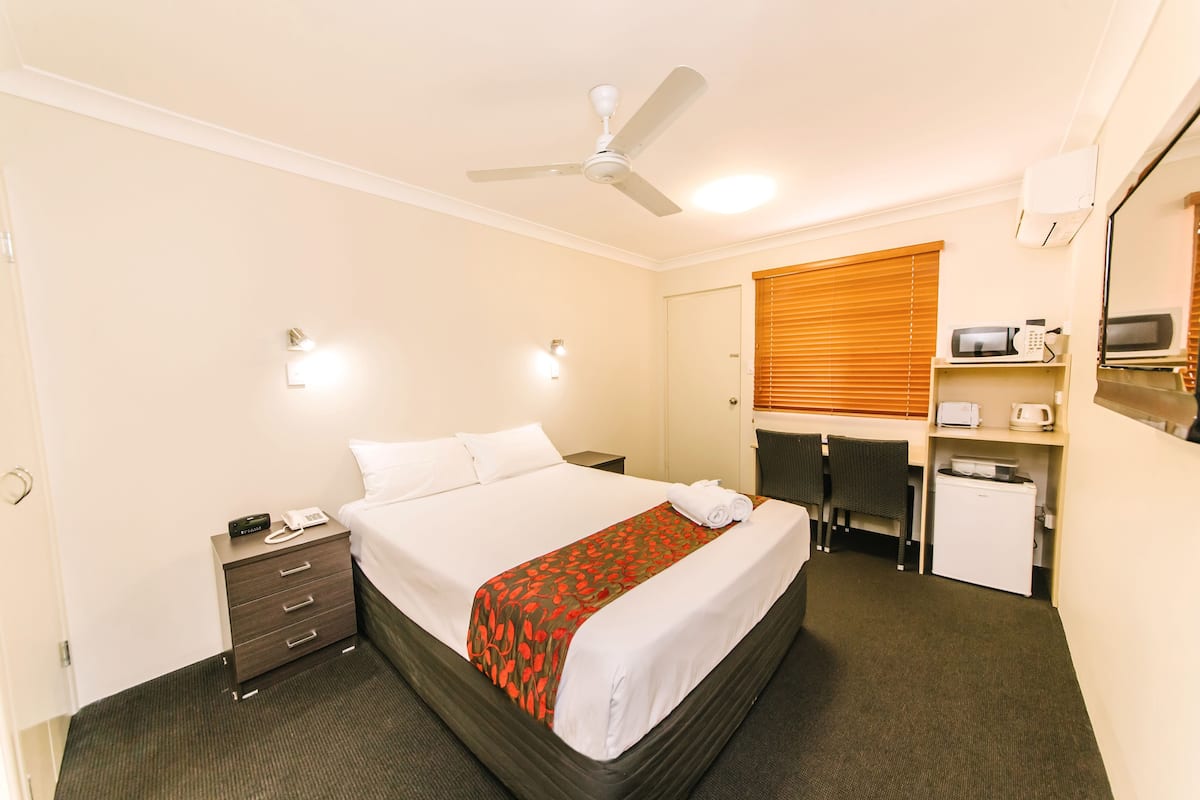 Habitación de hotel con cama, escritorio, silla, microondas, refrigerador, ventilador de techo y mesita de noche.