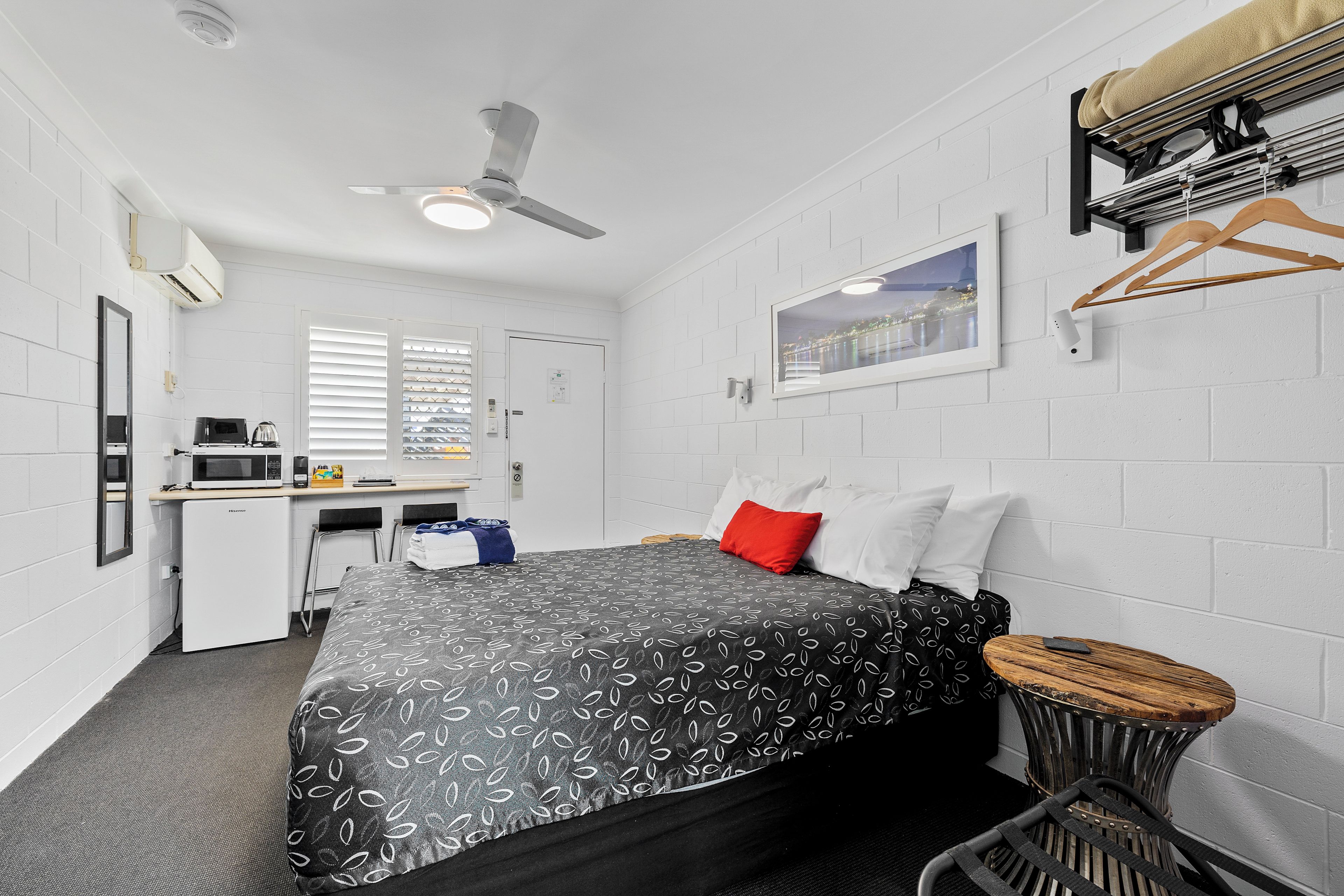 A hotel room with a bed, a desk, a chair, a hanging coat rack, and a window with blinds.