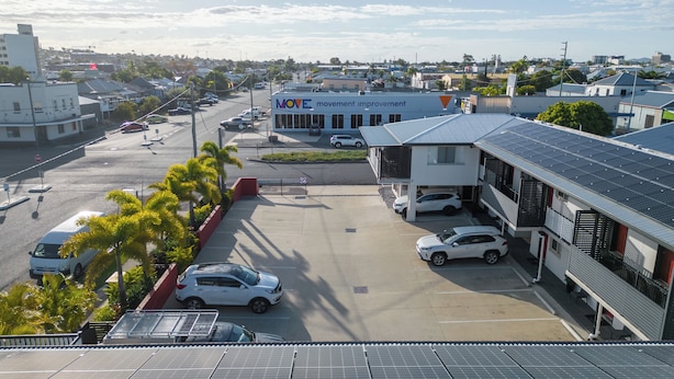 A parking lot with several cars, a building labeled "MOVE" with solar panels, and a view of a residential area with houses and palm trees. - Citywalk Motor Inn (Rockhampton)