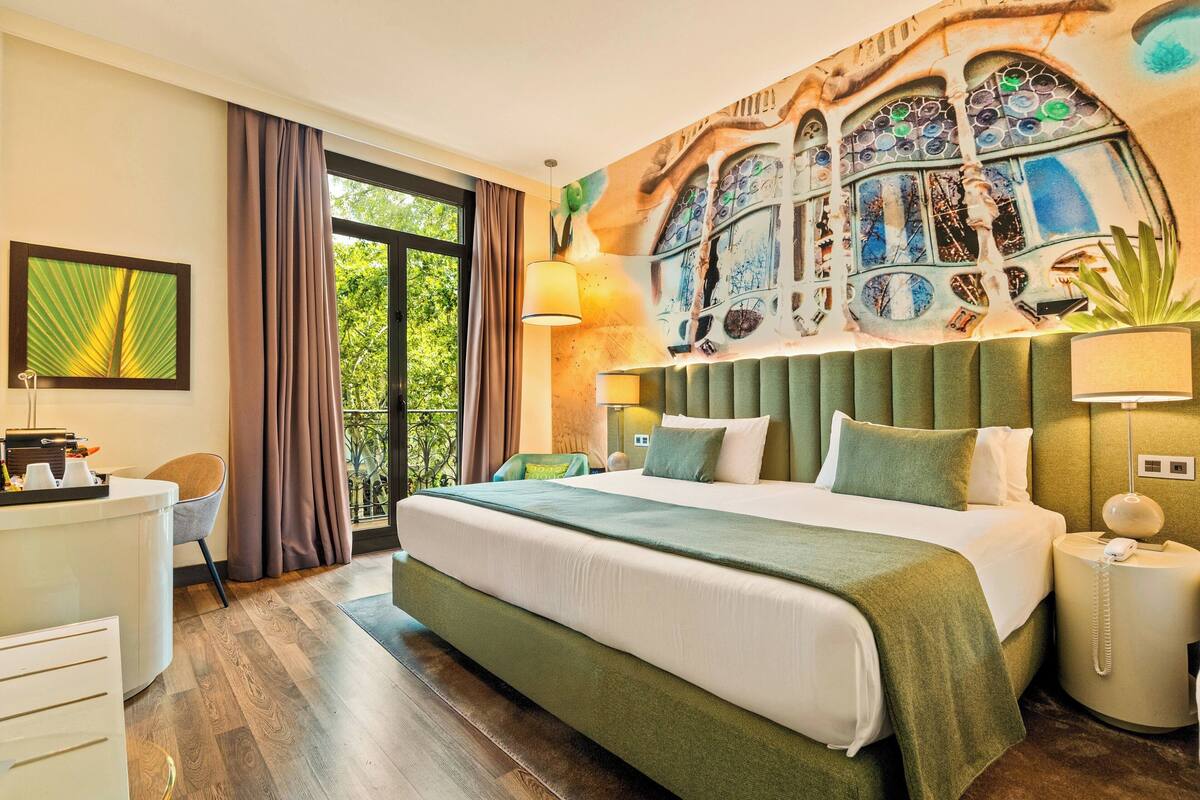 Ein Hotelzimmer mit einem großen Bett, einem Schreibtisch, einem Stuhl, einem Fenster mit Vorhängen und einem Wandgemälde.