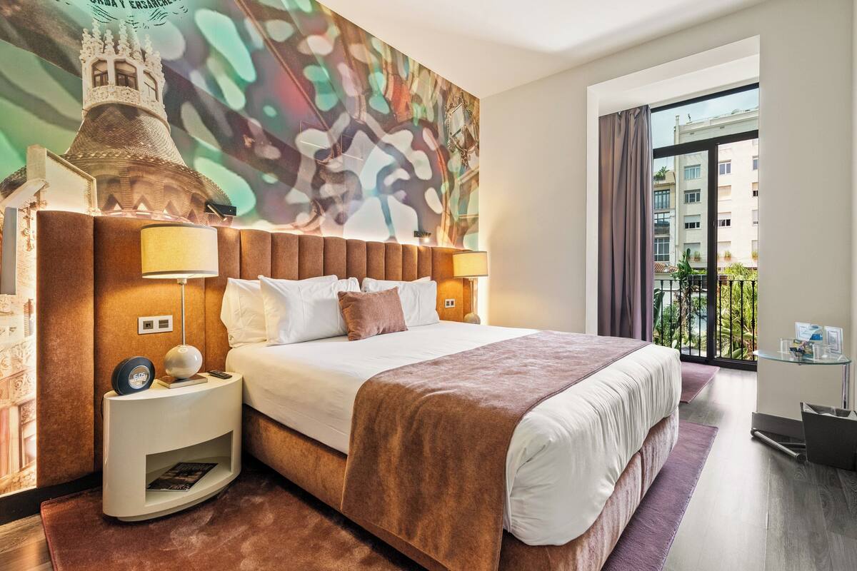 Ein modernes Hotelzimmer mit einem großen Bett, einem Nachttisch mit Lampe, einer Wanduhr und Blick auf die Stadt durch ein großes Fenster.