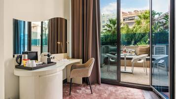 A hotel room with a curved white desk, a beige chair, a large sliding glass door leading to an outdoor patio with a table and chairs.