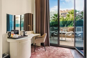 Une chambre d’hôtel avec un bureau blanc aux lignes courbes, une chaise beige et une grande porte-fenêtre coulissante donnant sur une terrasse extérieure avec une table et des chaises.