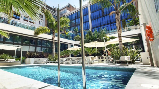 Ein Schwimmbecken mit Liegestühlen und Sonnenschirmen, umgeben von Palmen und modernen Gebäuden.