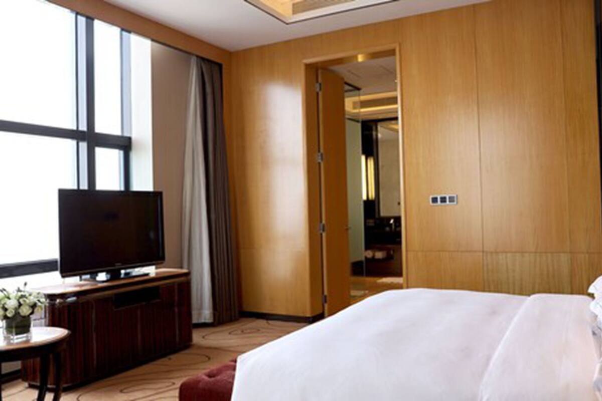 Habitación de hotel con una cama grande, un televisor sobre un mueble de madera, una mesita con un florero y una puerta que da al baño.