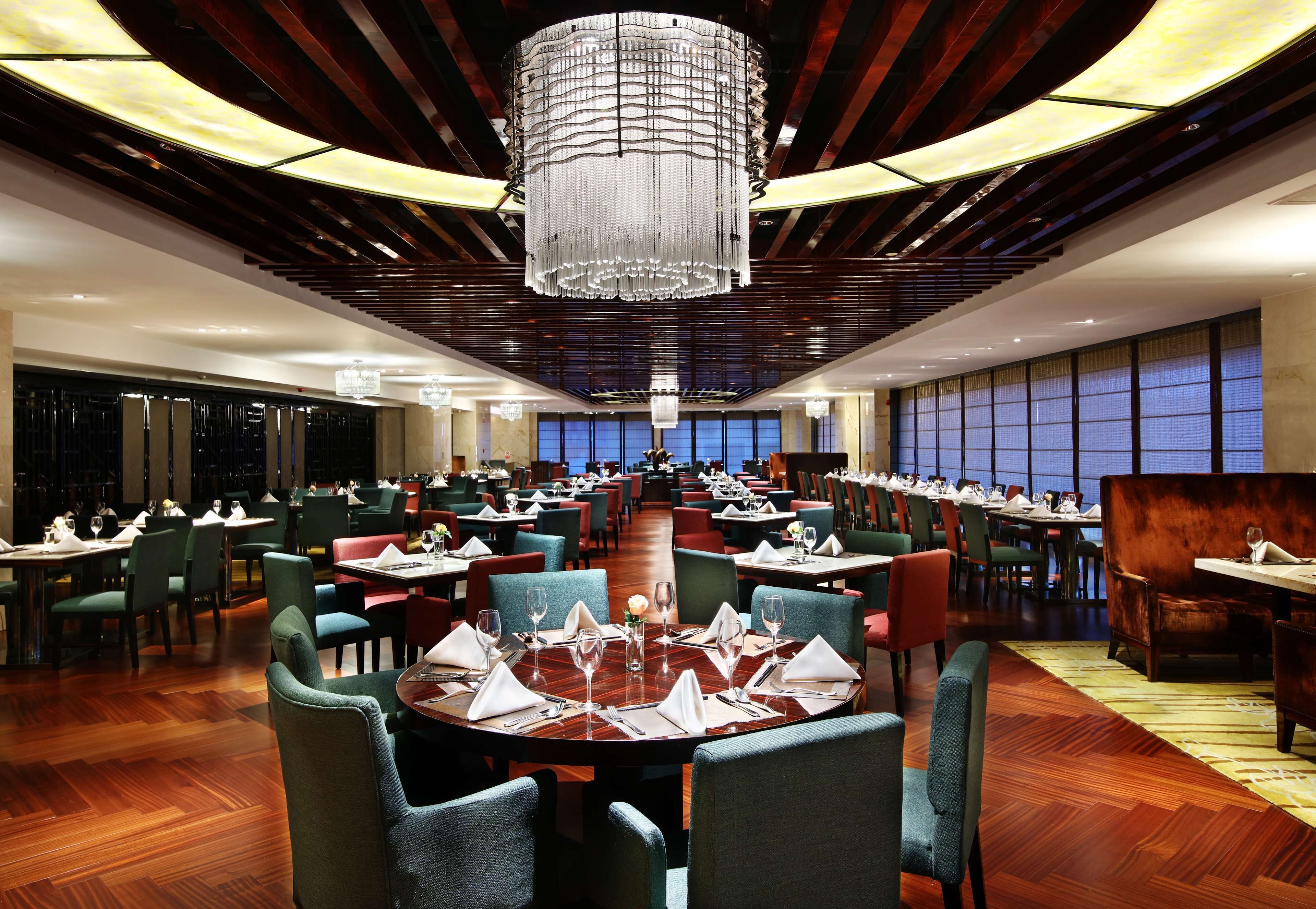 A spacious hotel dining area with multiple tables set for dining, a large chandelier, and a view of the cityscape through large windows.
