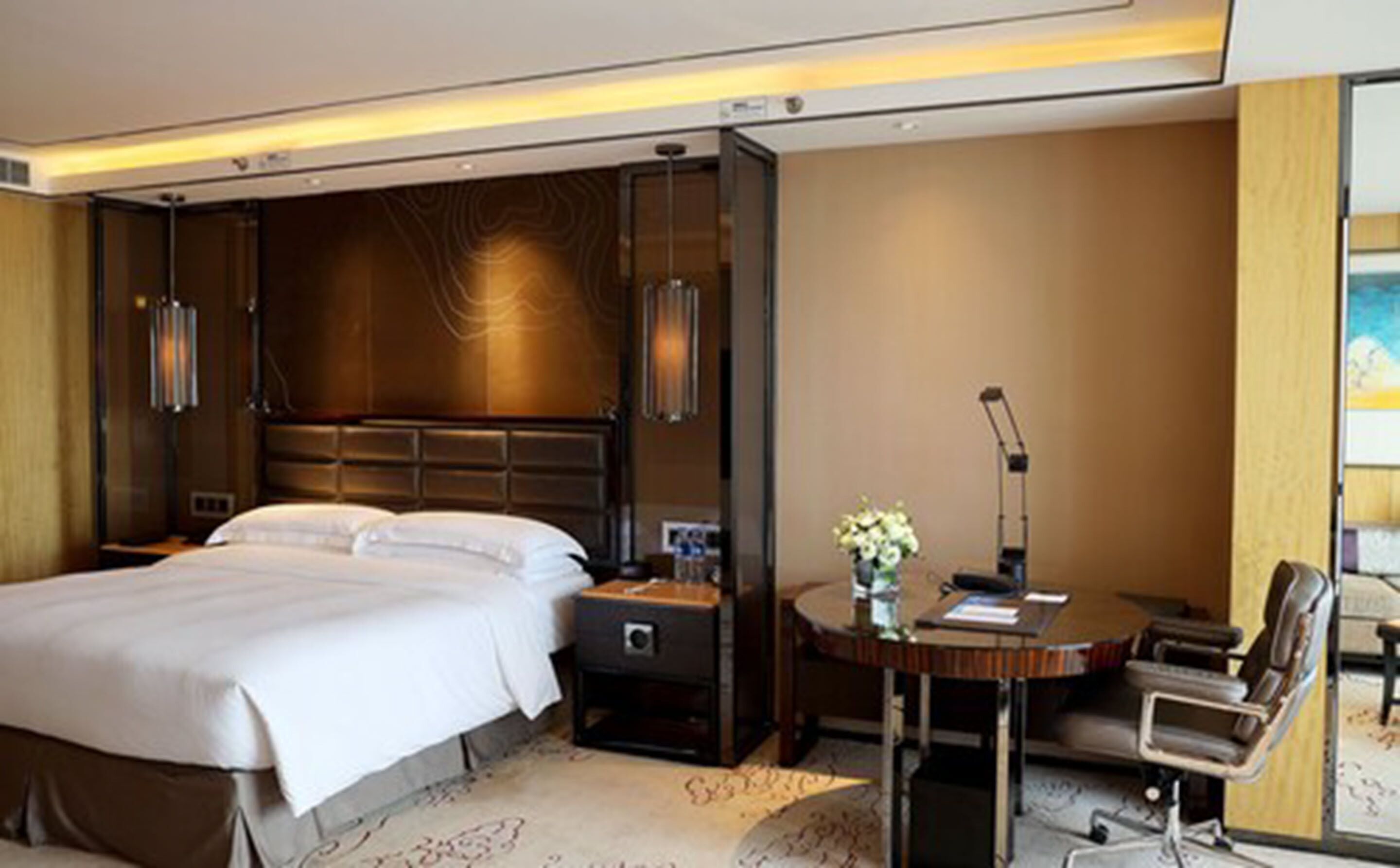 A hotel room with a large bed, a desk with a chair, a lamp, and a vase with flowers.