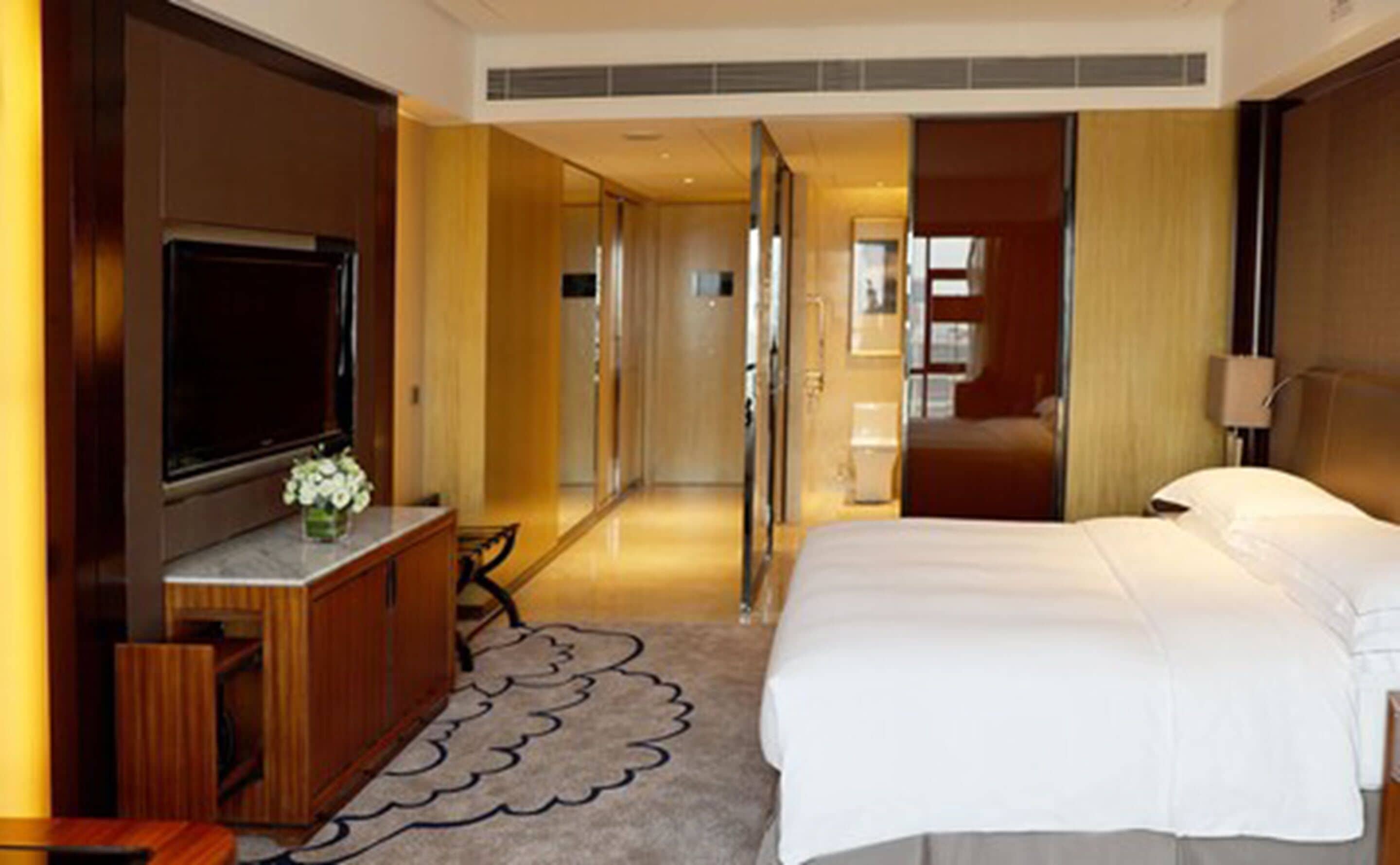 A hotel room with a large bed, a TV, a desk with a chair, and a bathroom visible through an open door.
