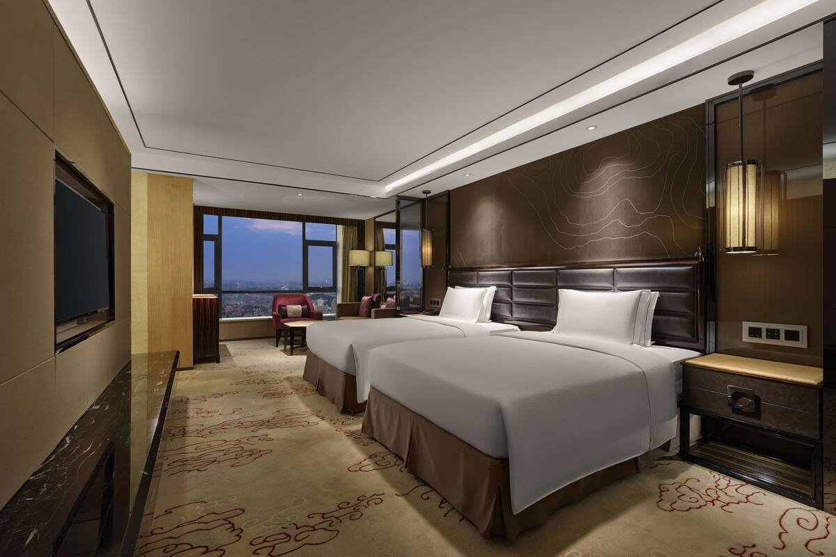 Habitación de hotel con dos camas, un ventanal grande con vistas a la ciudad, un escritorio con una silla y un televisor.