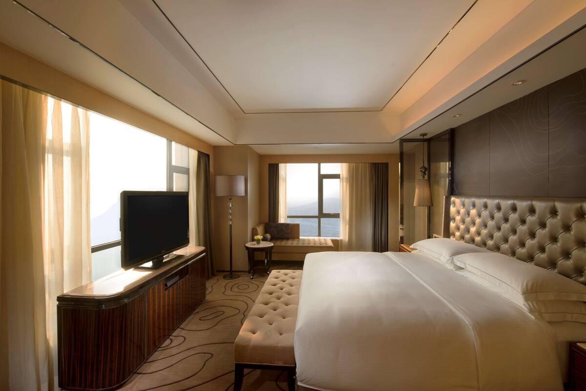 Una habitación de hotel con una cama grande, un televisor sobre una consola de madera, una mesita con una lámpara y vistas al mar a través de amplios ventanales.