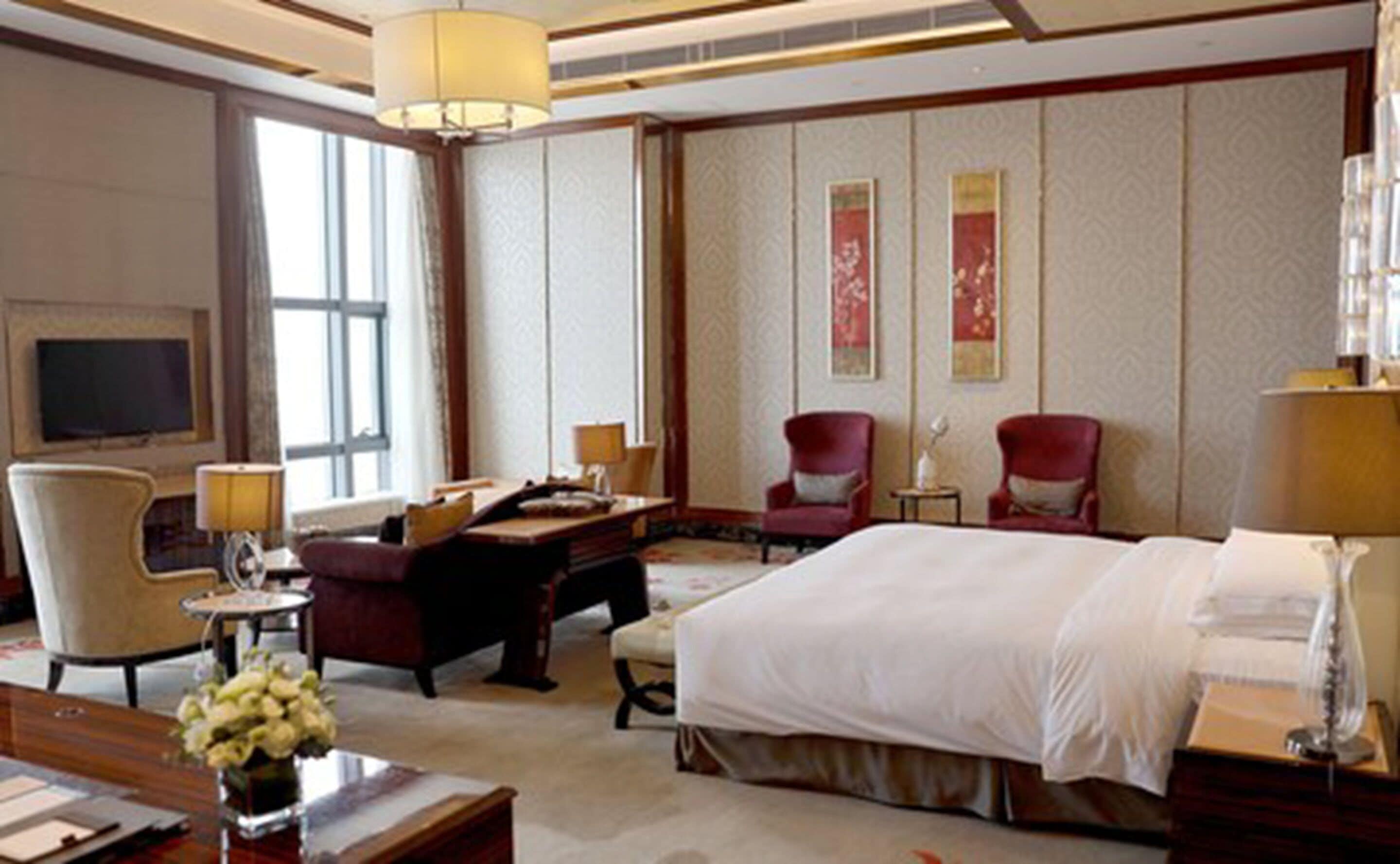 A hotel room with a large bed, two armchairs, a desk with a lamp, a TV, and decorative wall panels.