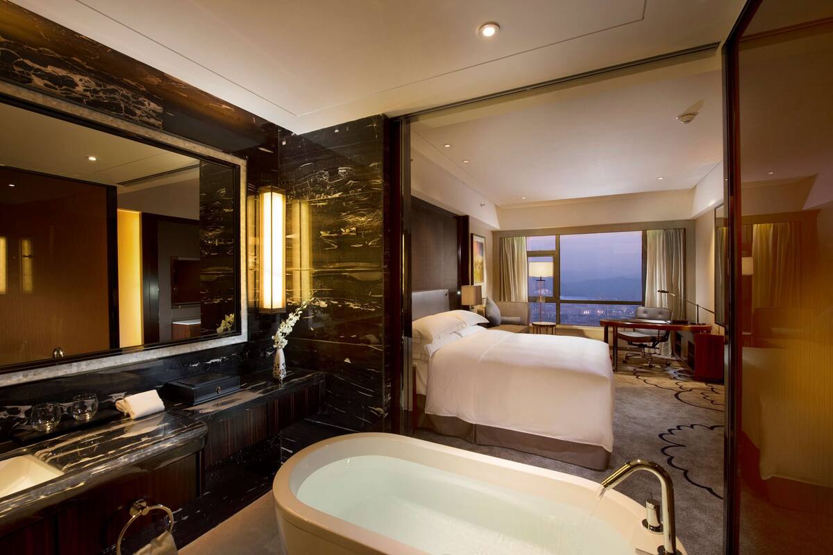 Una habitación de hotel moderna con una cama grande, una zona de estar con escritorio y silla, un ventanal con vistas y un baño separado con bañera y tocador.
