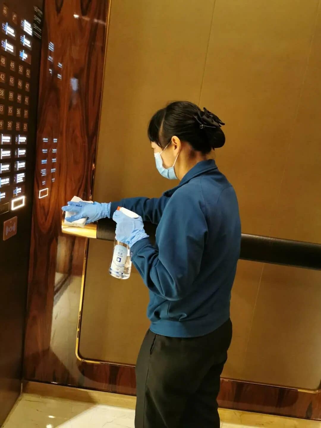 A person in a blue uniform cleaning an elevator with a spray bottle and cloth.