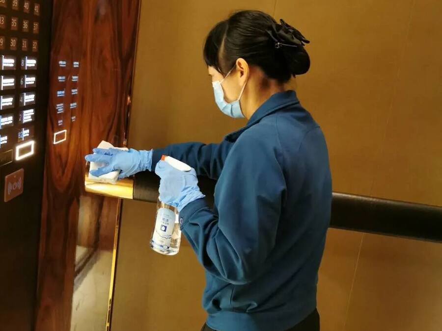 A person in a blue uniform cleaning an elevator with a spray bottle and cloth.
