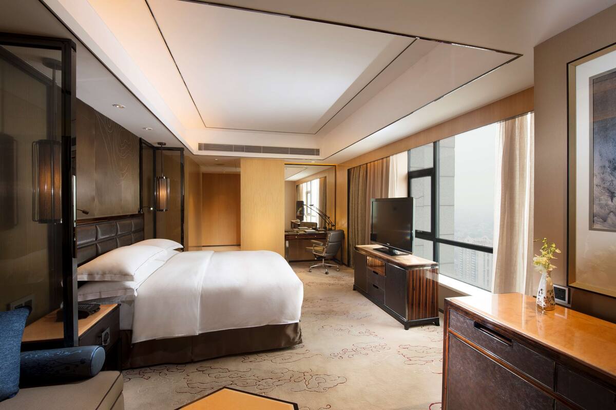 Una habitación de hotel moderna con una cama grande, un escritorio, un televisor y un ventanal amplio.