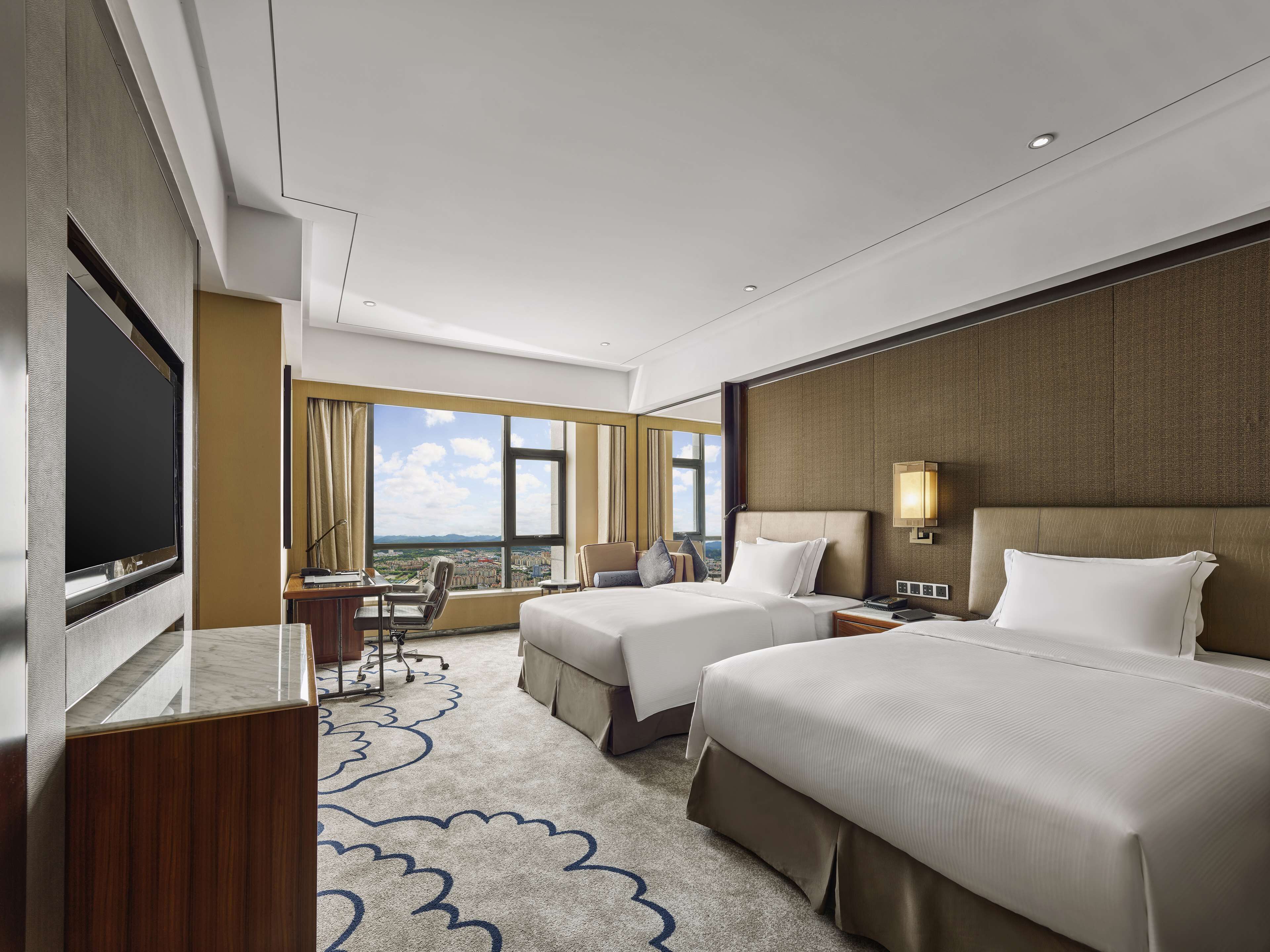Habitación de hotel con dos camas, un escritorio, un televisor y un ventanal con vistas a la ciudad.
