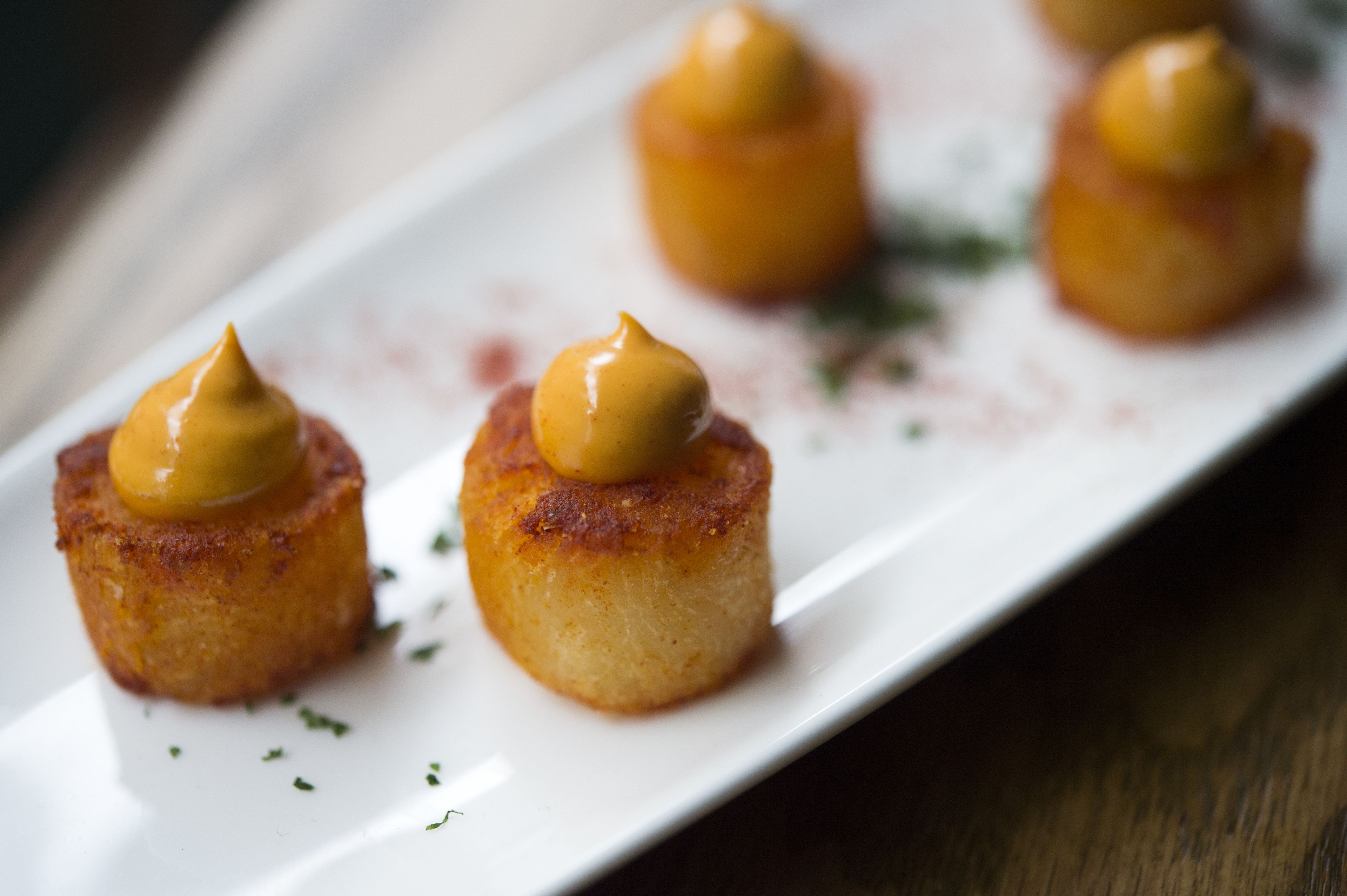Un plato blanco rectangular con tres pequeñas piezas redondas de color dorado oscuro, cubiertas con una cucharada de salsa amarilla, adornado con hierbas verdes.