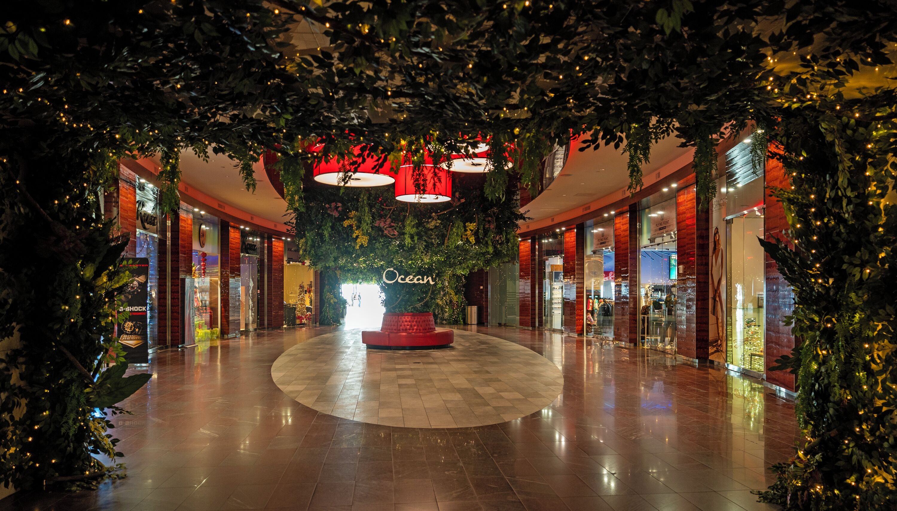 A mall interior with a central circular seating area, glass storefronts, and hanging lights.