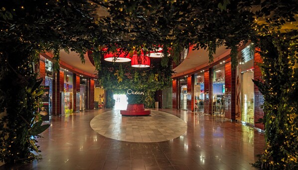 Interior de centro comercial con una zona de estar circular central, escaparates de vidrio y luces colgantes.