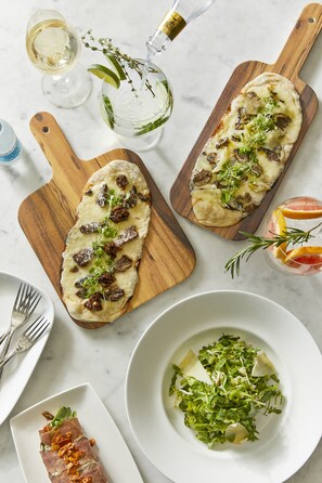 Deux planches à découper en bois, sur lesquelles sont disposées des galettes garnies de champignons et de légumes verts, un verre de vin blanc, un verre contenant une tranche d’agrumes et une branche de romarin, et une assiette de légumes verts accompagnée de copeaux de fromage.