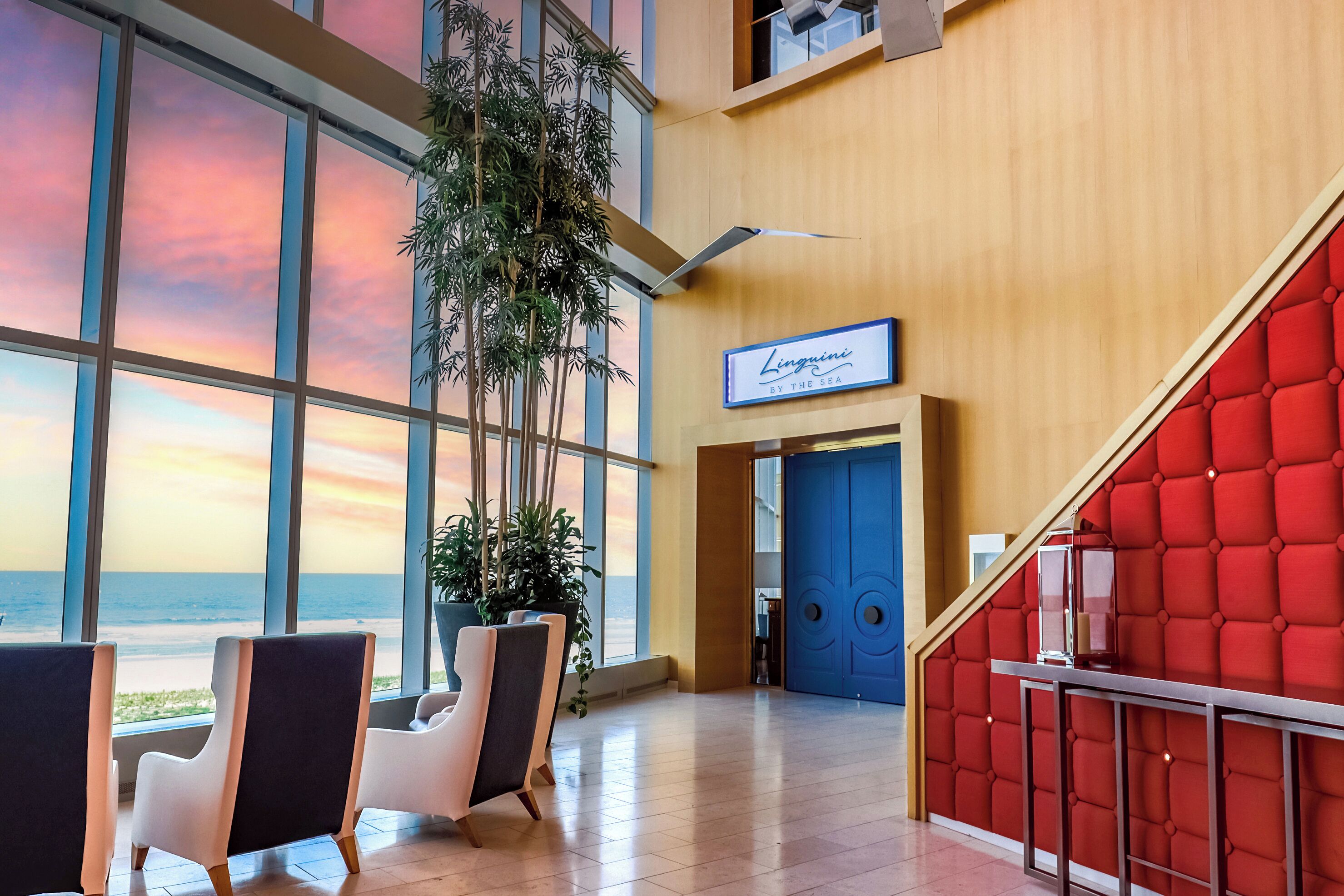 A hotel lobby with large windows, a staircase with red upholstery, and a sign reading 'Lingqun By The Sea'.