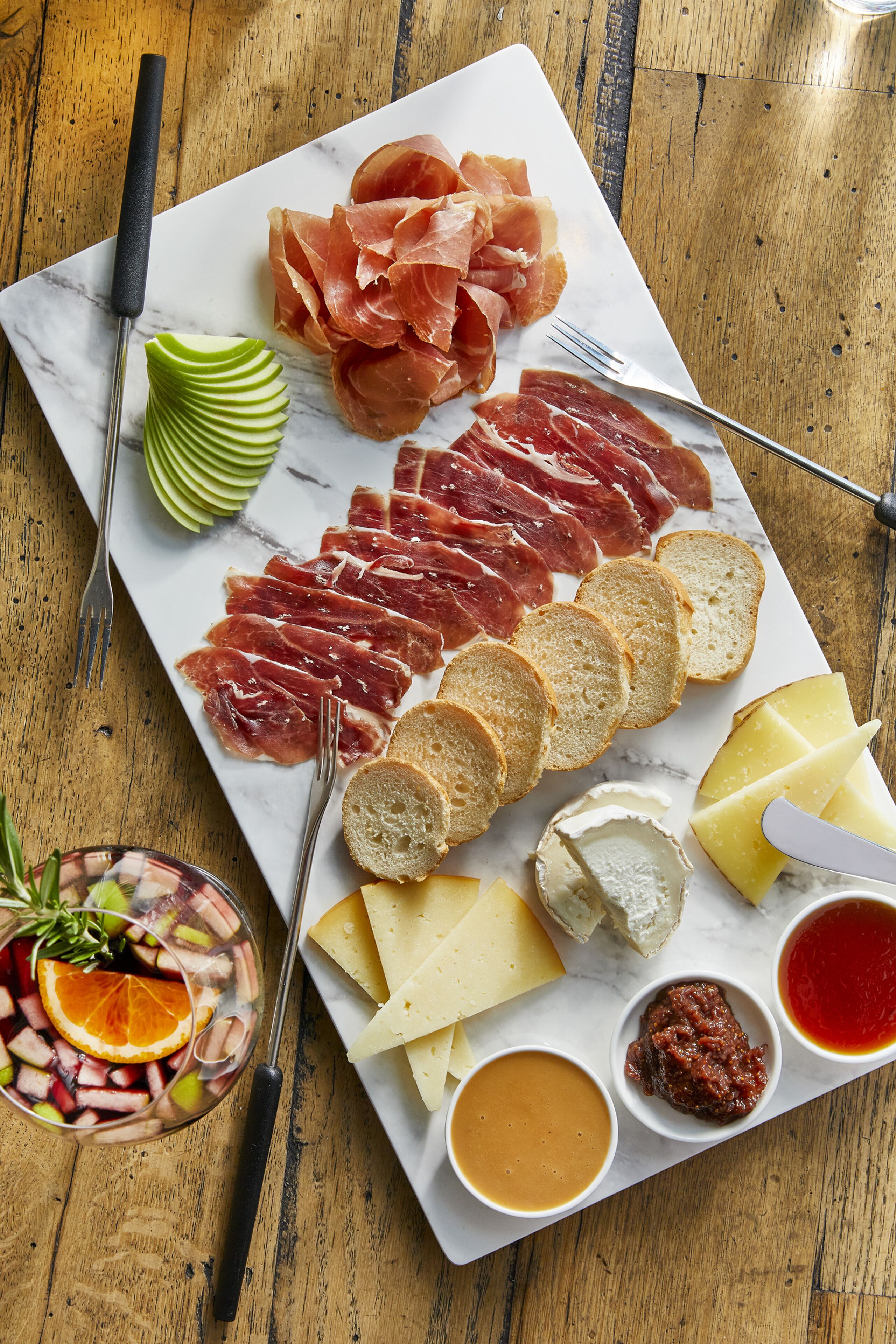 Una tabla de embutidos con diferentes carnes, quesos, pan y salsas sobre una superficie de mármol.