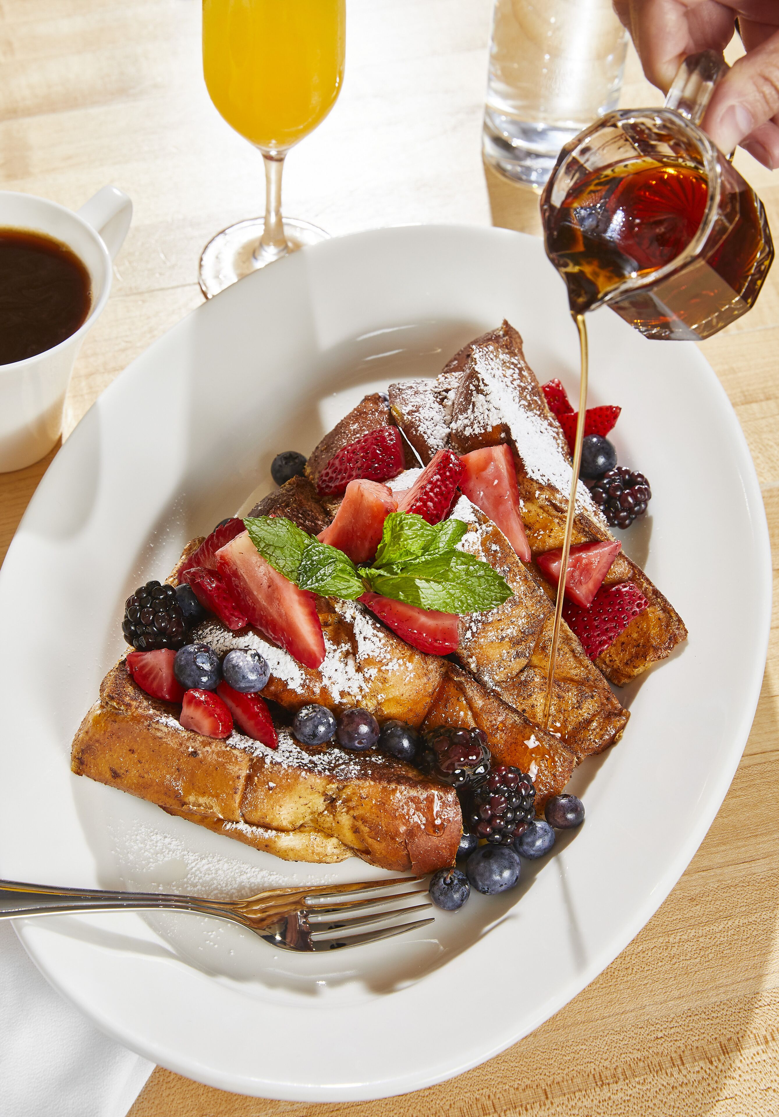 A plate of French toast with strawberries, blueberries, and blackberries, topped with powdered sugar and a mint leaf.