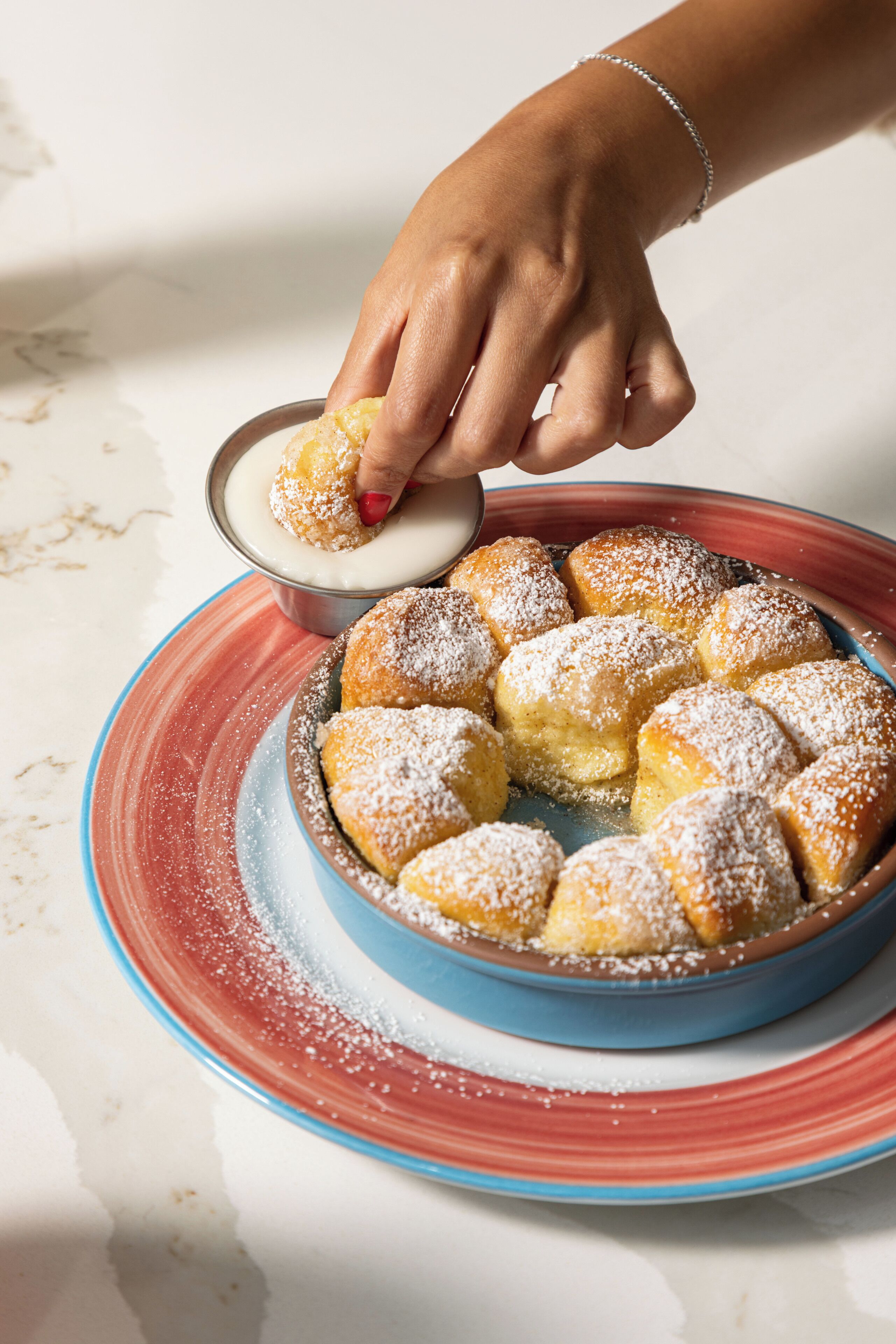 A hand dipping a doughnut into a cup of liquid, with a plate of doughnuts in the background.