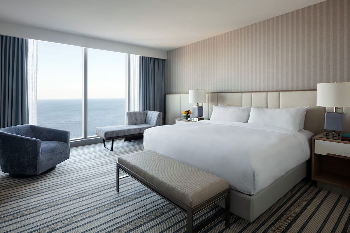 Una camera d'albergo con un letto grande, una sedia, una panca, un comodino e vista sull'oceano.