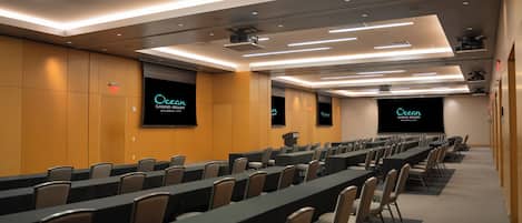 Un salón de conferencias con filas de mesas y sillas, una pantalla de proyector y una pantalla grande que muestra el nombre del hotel.