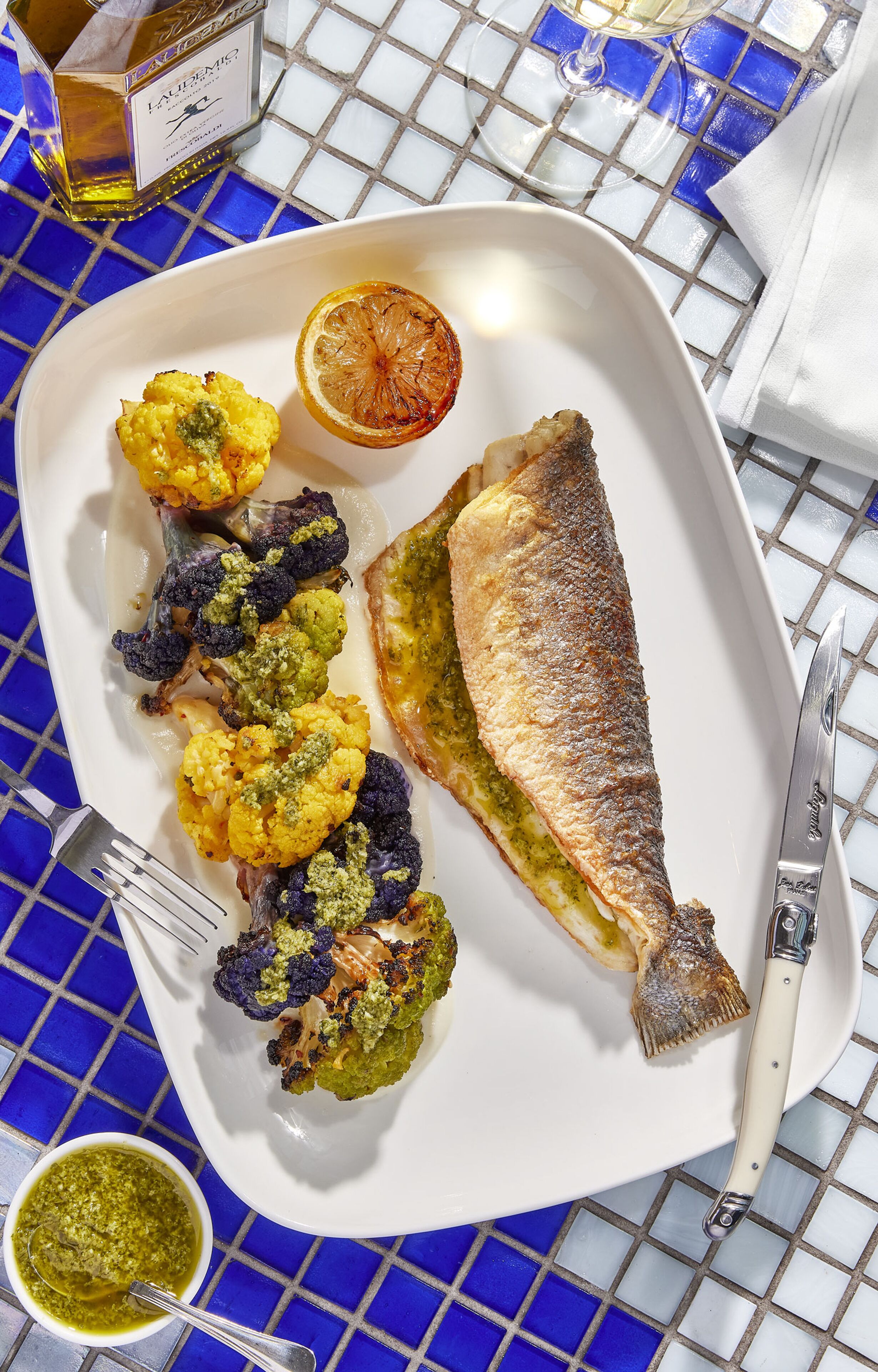 En tallrik med grillad fisk, rostad blomkål och en citronskiva, placerad på ett bord med kakelplattor.